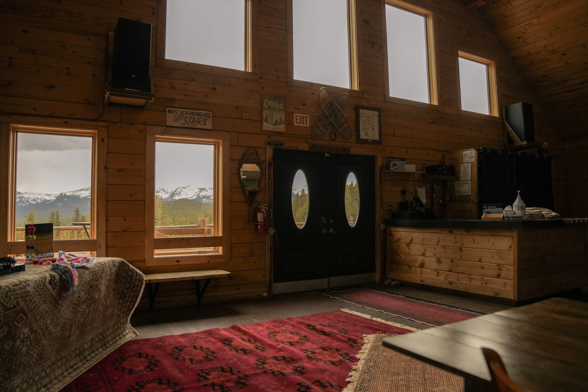 Interior of a wooden cabin with large windows showing mountain scenery, with signs on the wall, a black double door, and a patterned red and beige rug on the floor.