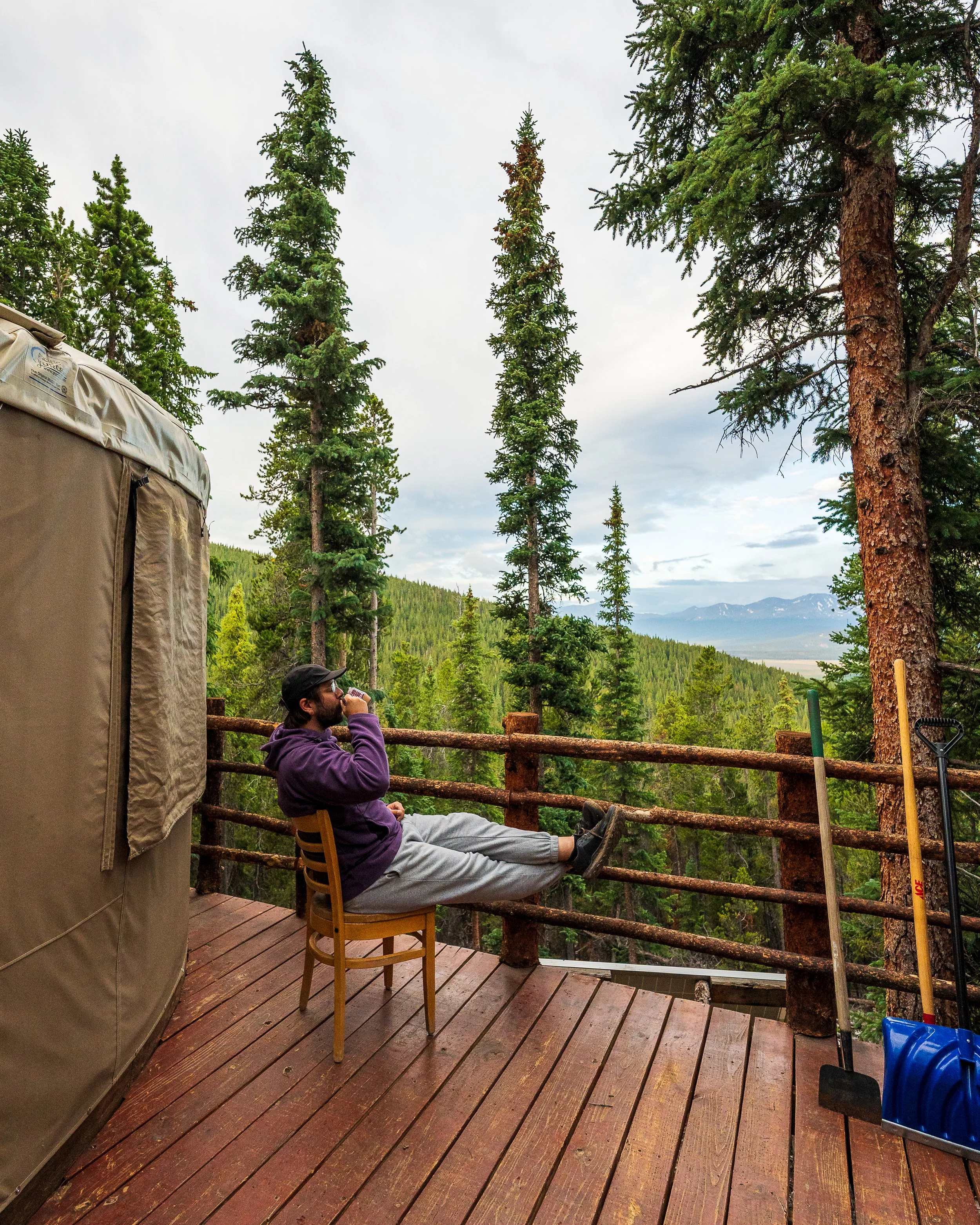 A person with dark hair, sunglasses, and a purple hoodie is sitting on a wooden deck, relaxing and drinking from a mug while overlooking a scenic view of trees, hills, and mountains under a cloudy sky.
