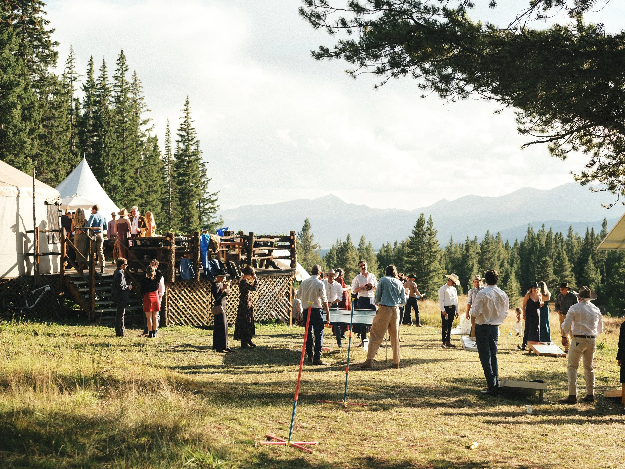 A group of people gathered outdoors in a mountainous forest area with tents and trees, participating in a recreational activity on a sunny day.