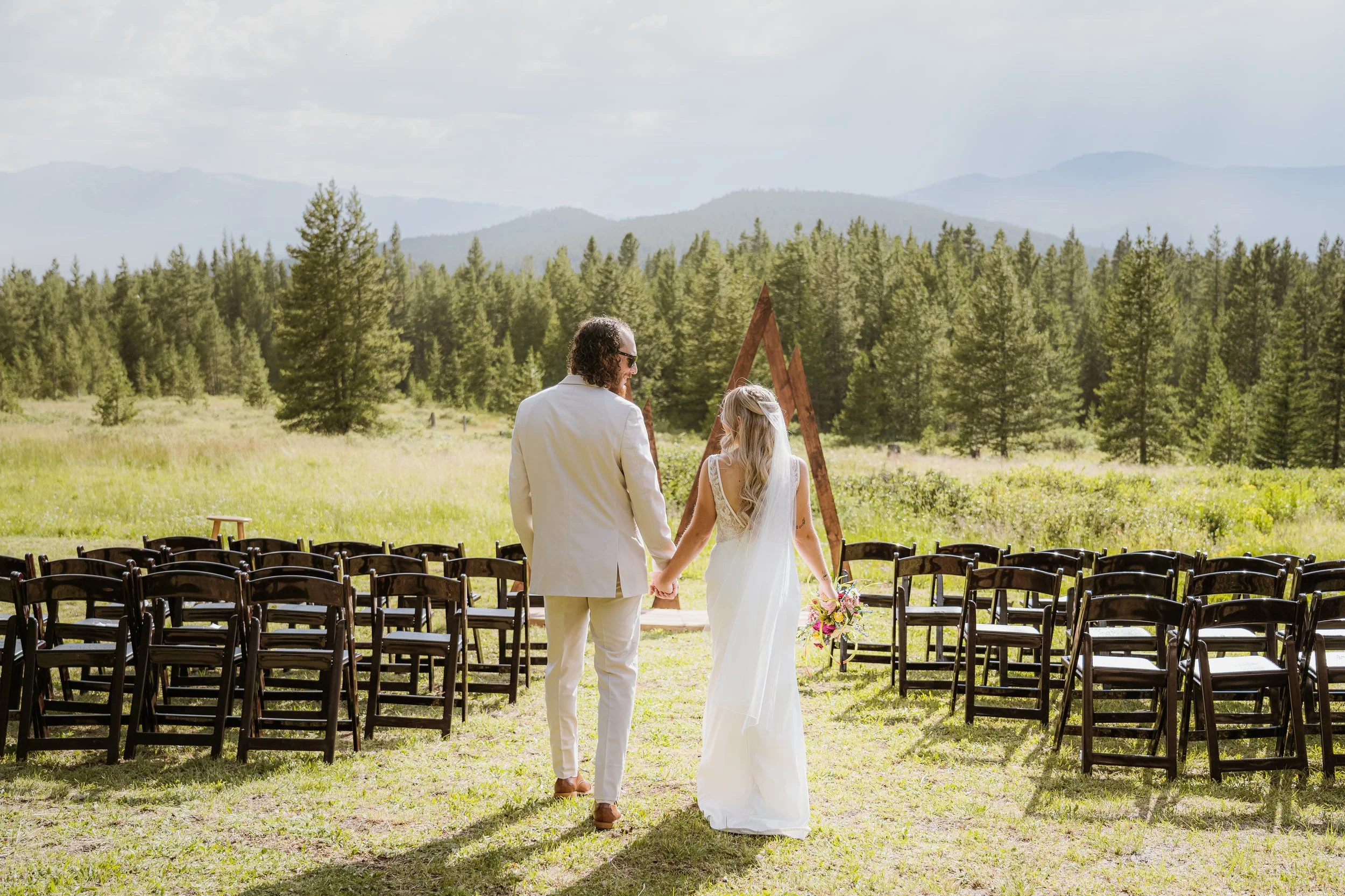 A bride and groom holding hands and walking towards an outdoor wedding altar in a lush green meadow with mountains in the background.