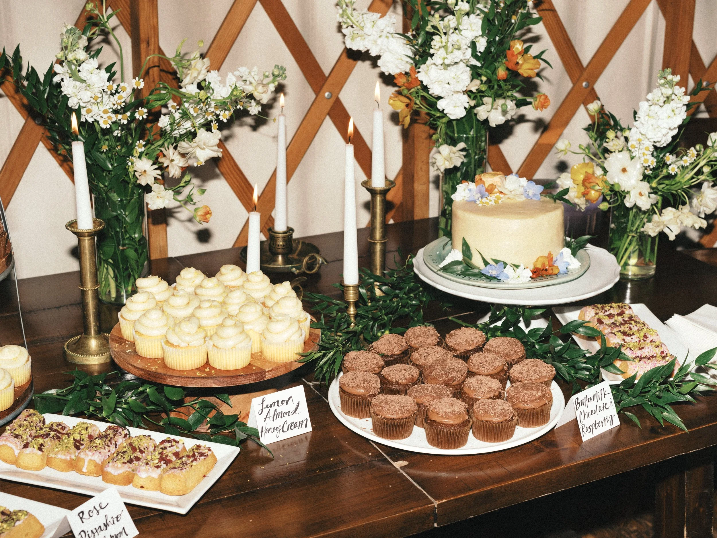 Dessert table with various treats including lemon almond honey cupcakes, chocolate raspberry cupcakes, and cookies, surrounded by floral arrangements with white, yellow, and purple flowers and candles.