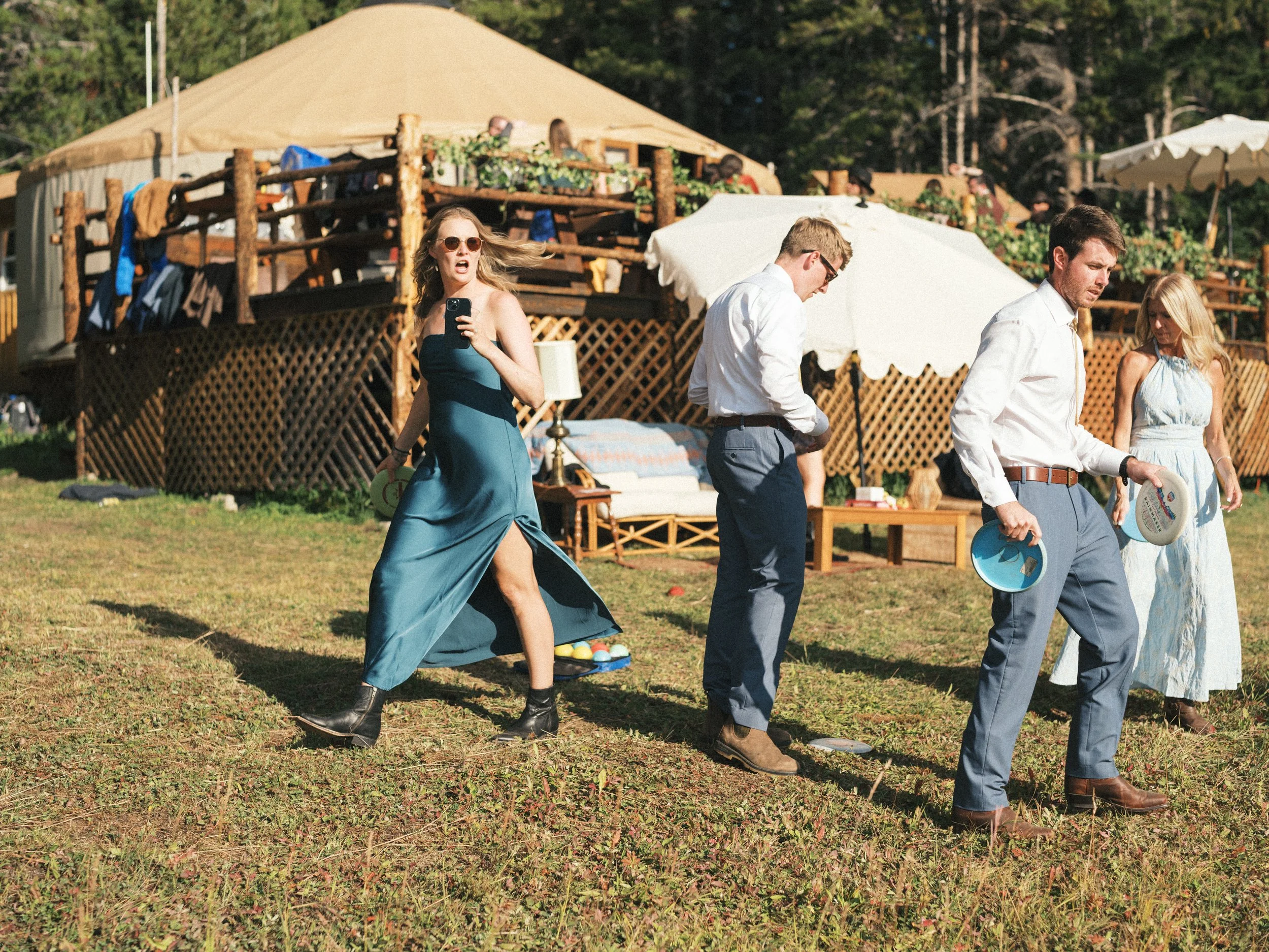 Group of people outdoors playing disc golf on a grassy field near a wooden deck and a tent, with trees in the background.