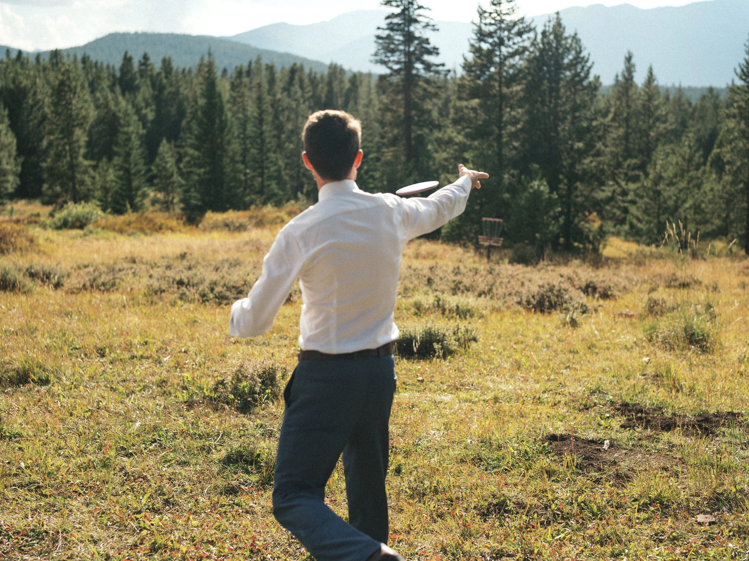 A man dressed in a white shirt and dark pants is standing outdoors in a grassy field surrounded by trees. He is facing away from the camera and pointing to the sky, with mountains visible in the background.