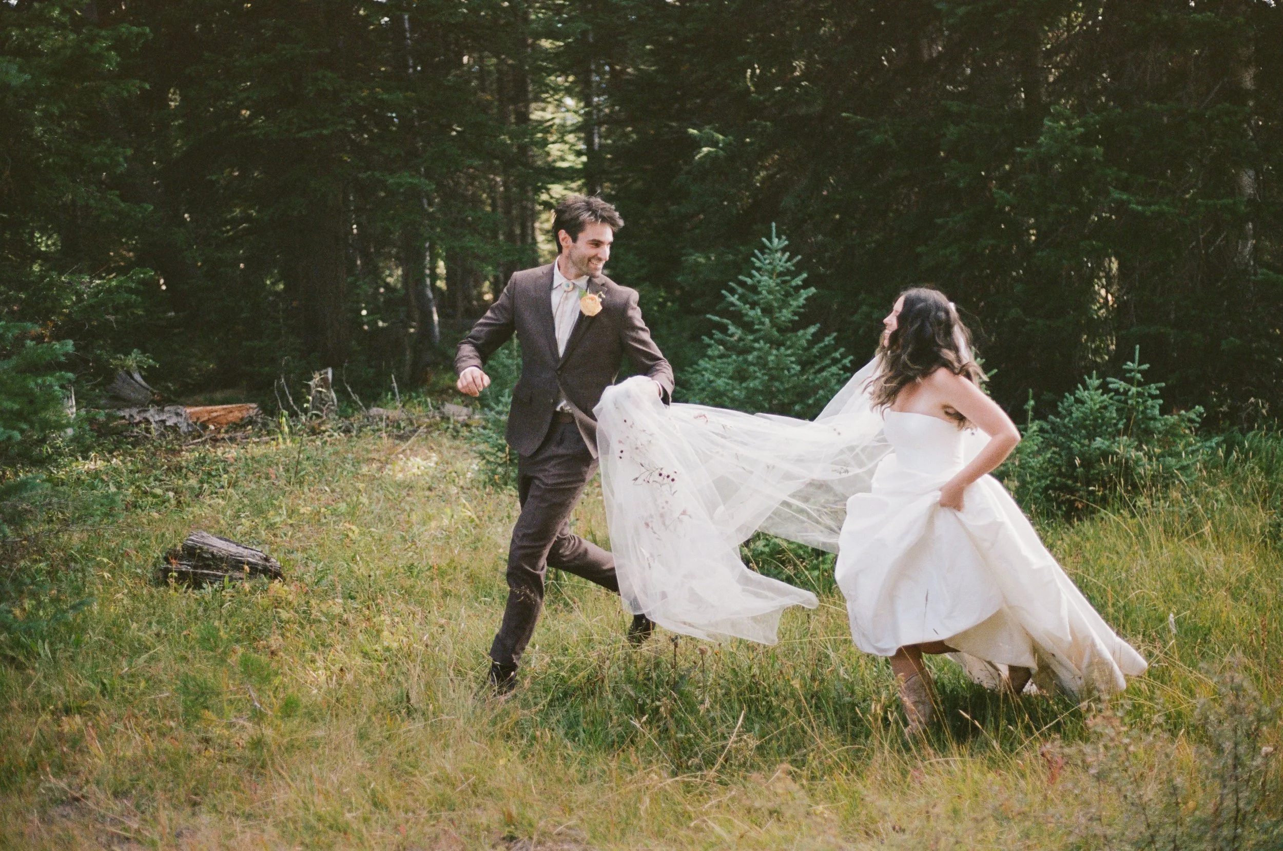 A man in a suit and a woman in a wedding dress dance in a forest with tall trees and green underbrush, creating a joyful scene.