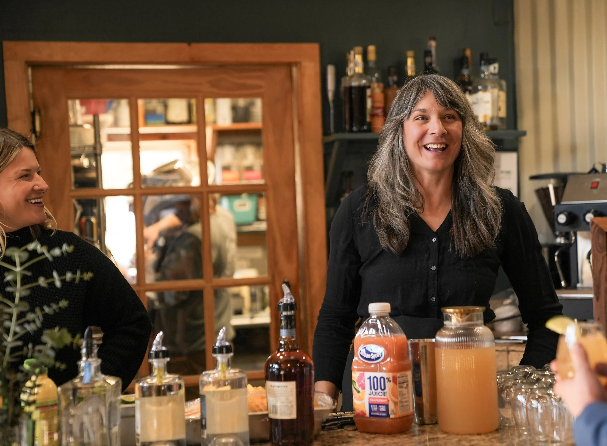 Two women smiling and talking in a cozy restaurant or cafe setting, with bottles and condiments on the table.