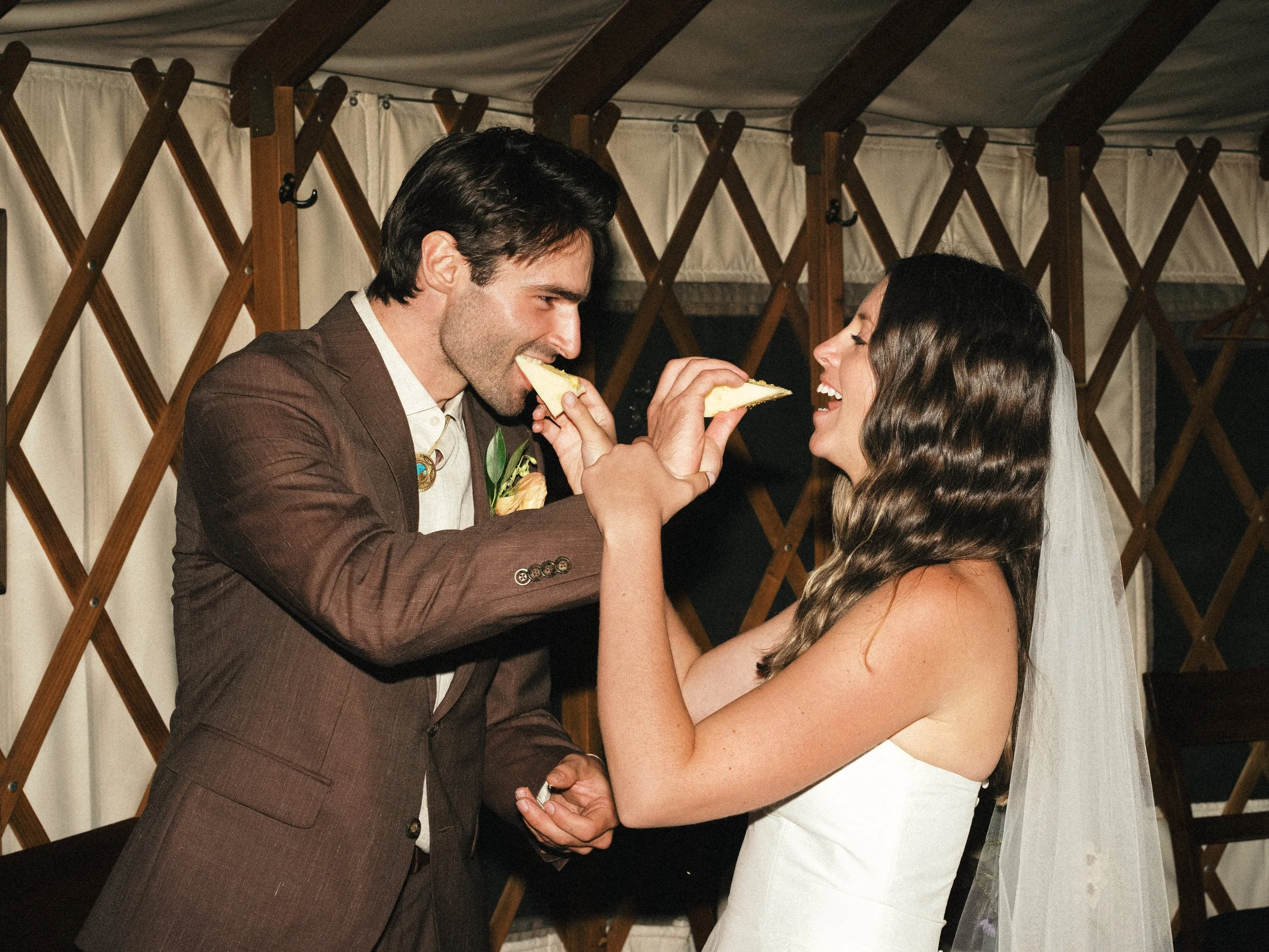 A man and woman, both dressed formally, sharing a moment of joy as the man feeds the woman a slice of pizza during a celebration or wedding.