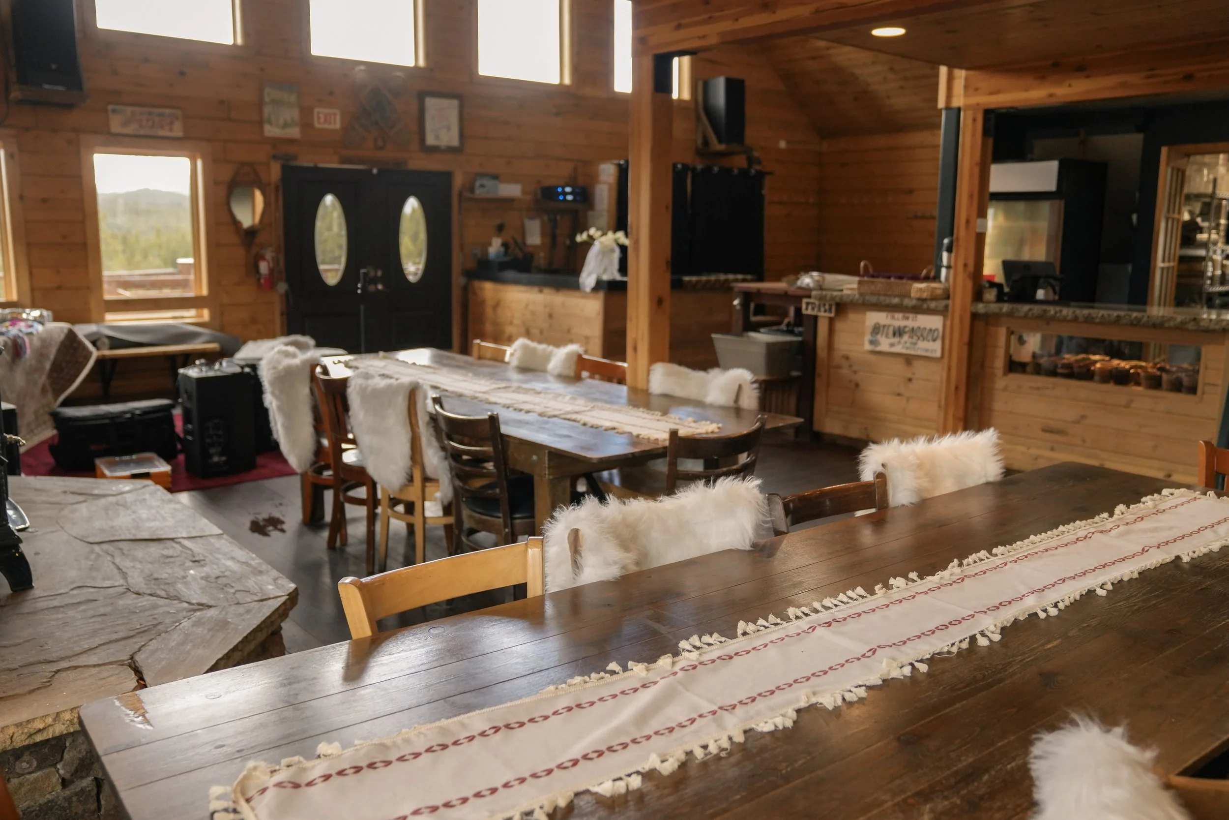 Rustic indoor space with wooden walls, featuring a dining area with a long wooden table covered with a decorative table runner and surrounded by chairs with fluffy white throws. In the background, there is a kitchen area with a granite countertop, sh