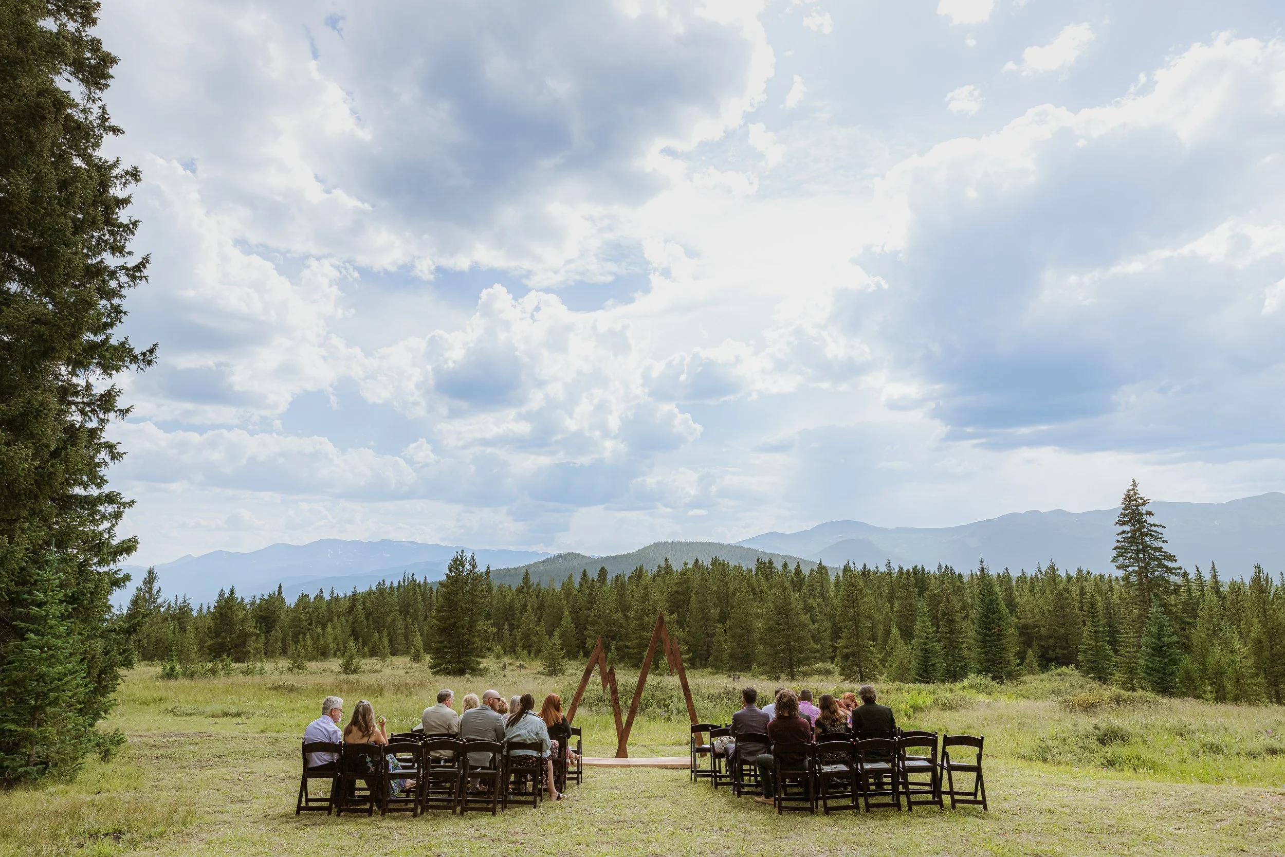 An outdoor wedding ceremony taking place in a scenic mountain setting with forests and mountains in the background, and a group of people sitting on chairs in front.