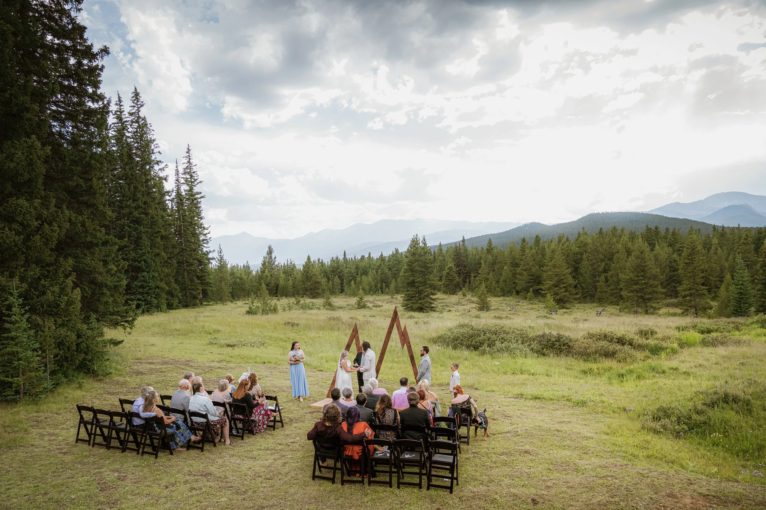 A wedding ceremony outdoors on a grassy field surrounded by tall trees and mountains in the background. Brides and grooms stand under a wooden arch, exchanging vows. Guests sit on chairs and benches facing the couple. The sky is partly cloudy.