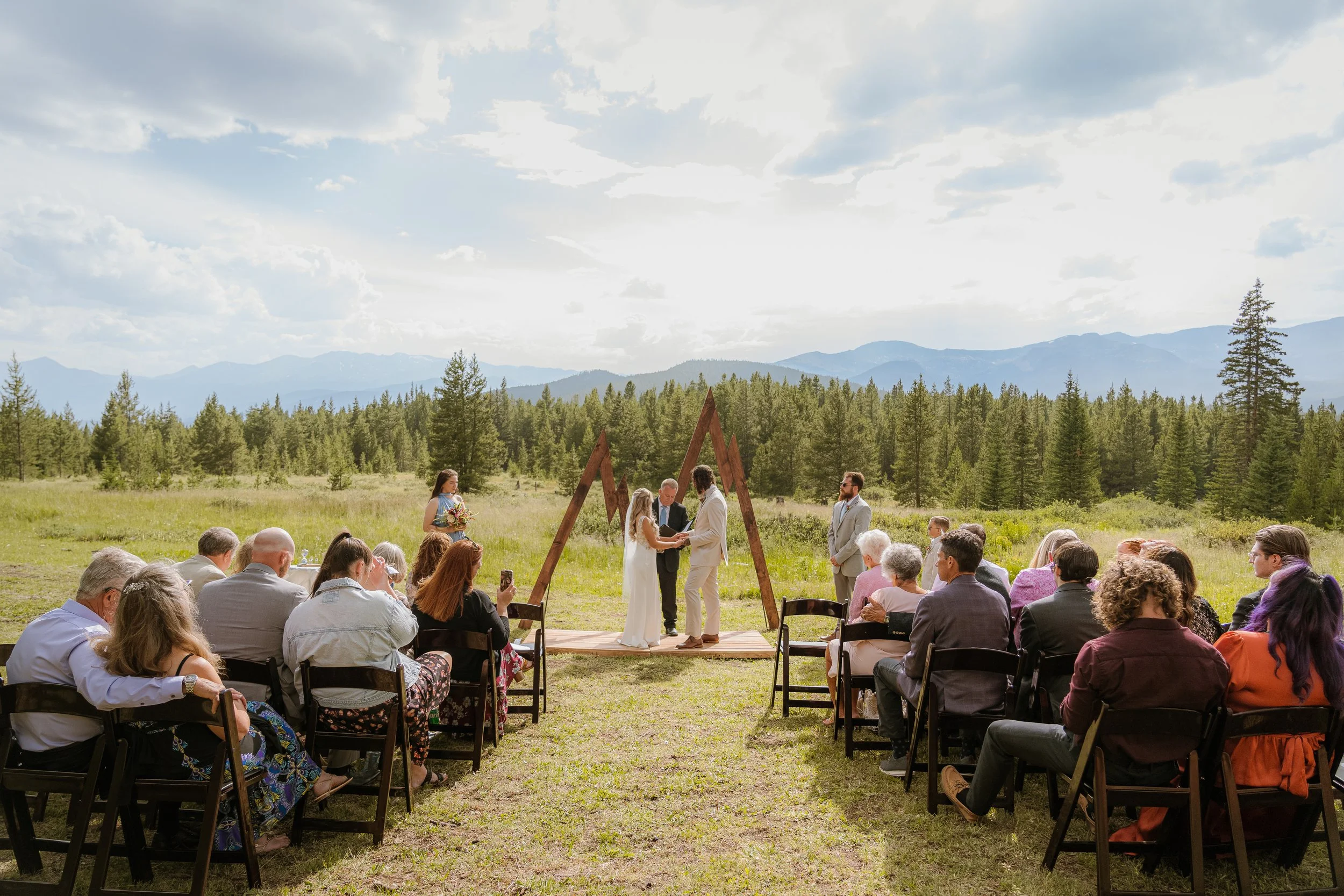 Outdoor wedding ceremony taking place in a meadow surrounded by trees and mountains, with guests seated and couple exchanging vows under a triangular wooden arch.