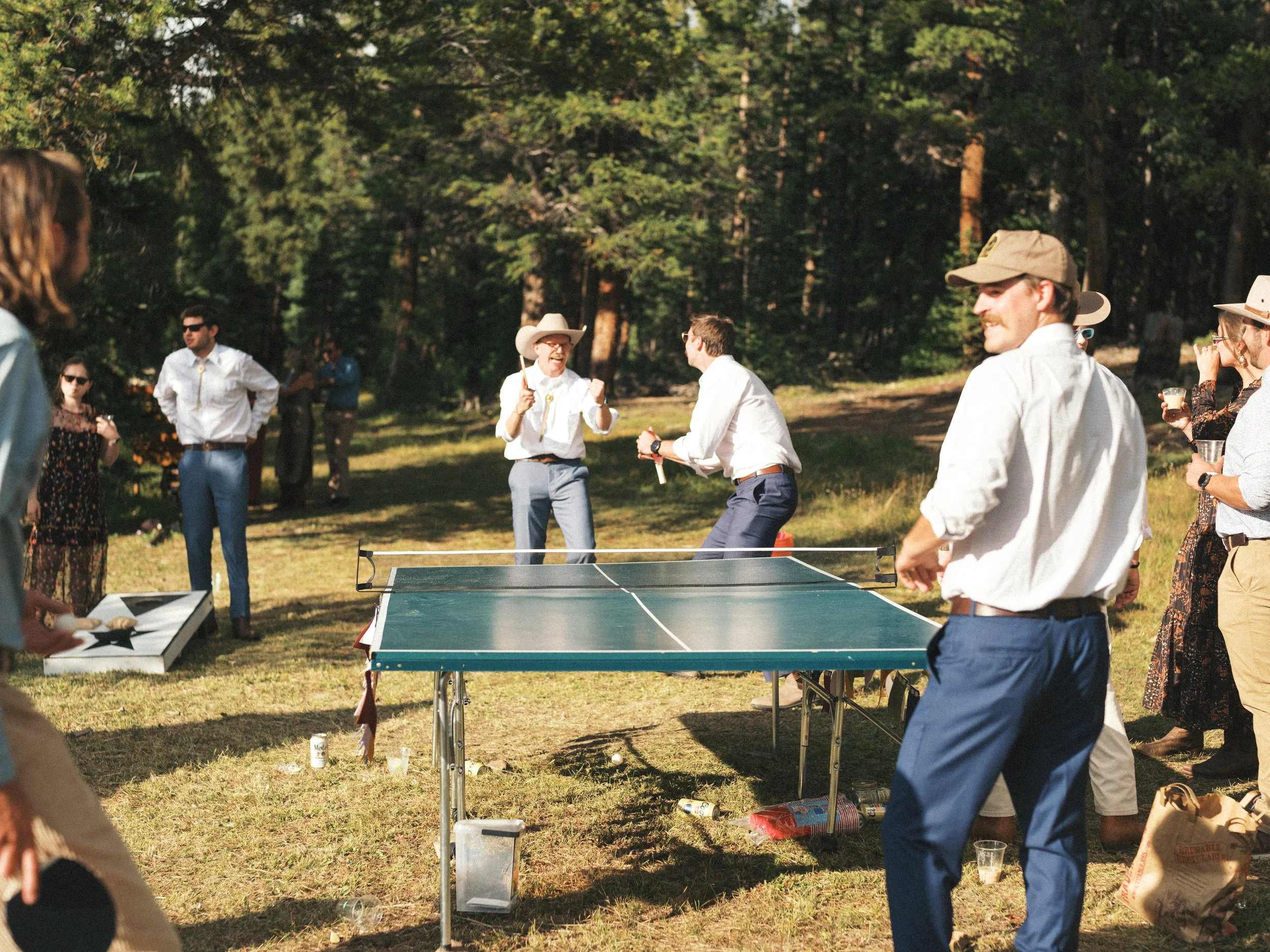 People playing ping pong outdoors at a gathering in a wooded area, with some holding drinks and wearing casual and semi-formal clothing, including hats and sunglasses.