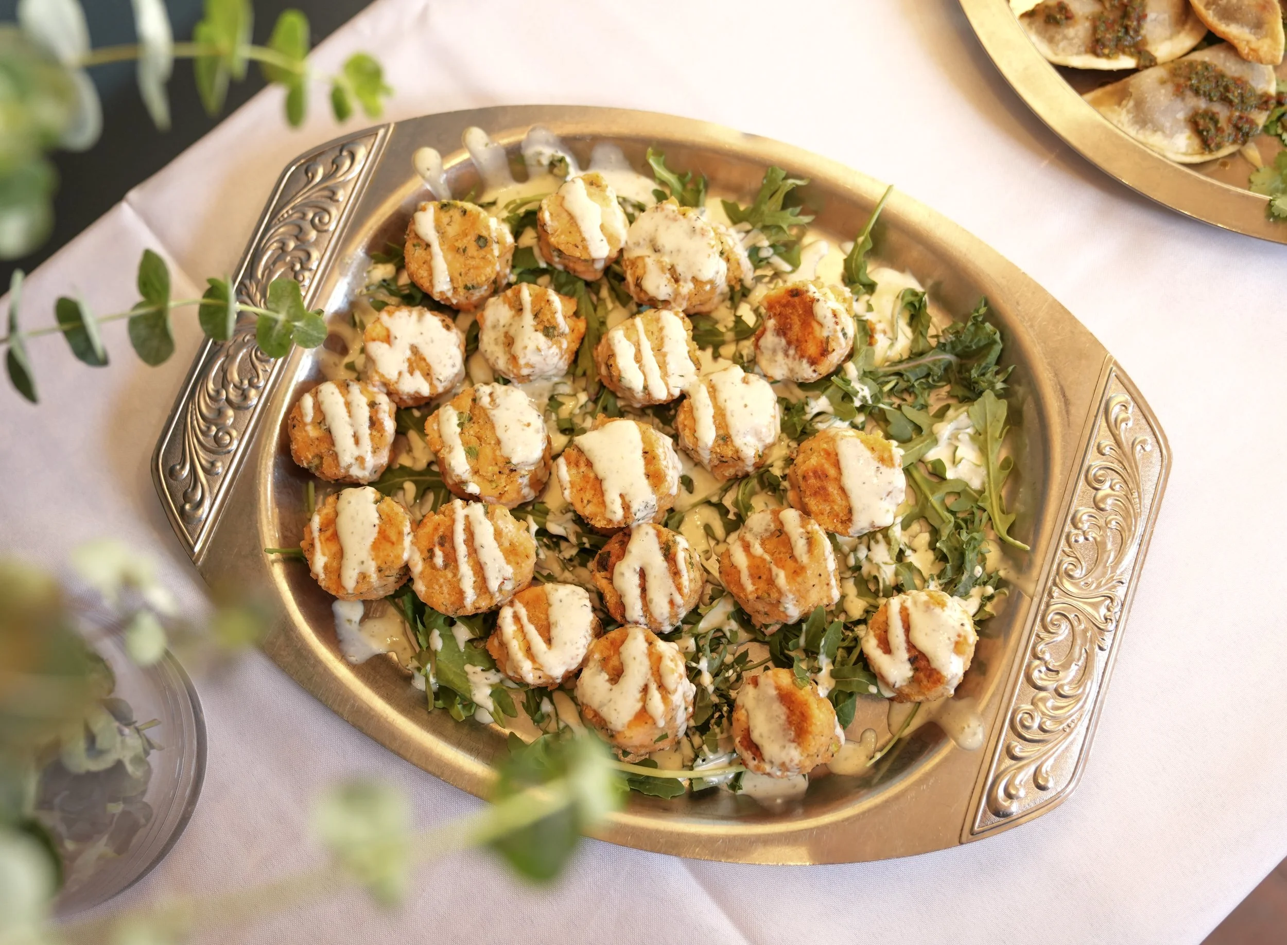 A silver tray filled with fried balls topped with white sauce, sitting on a white tablecloth with a side of crepes topped with green herbs and red sauce.