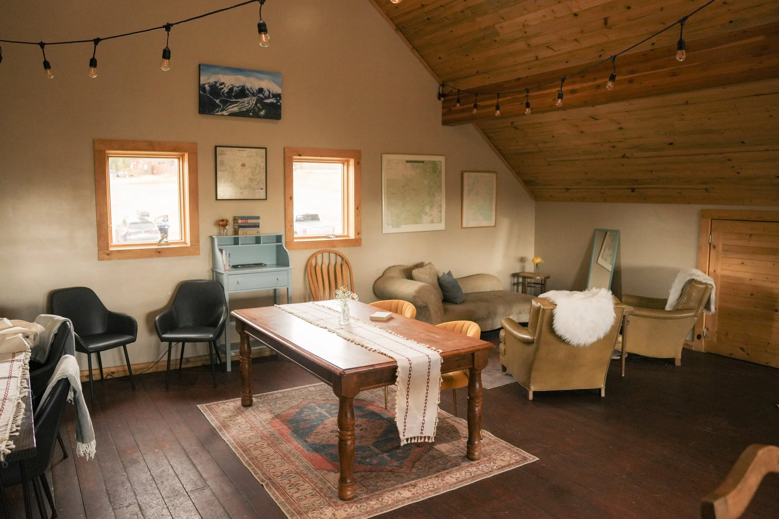 A cozy living room with wooden ceiling and trim around windows and door. It features a wooden table with a cloth runner and decorative items, a rug, multiple chairs, a sofa with cushions, and framed artwork on the walls. String lights hang across the