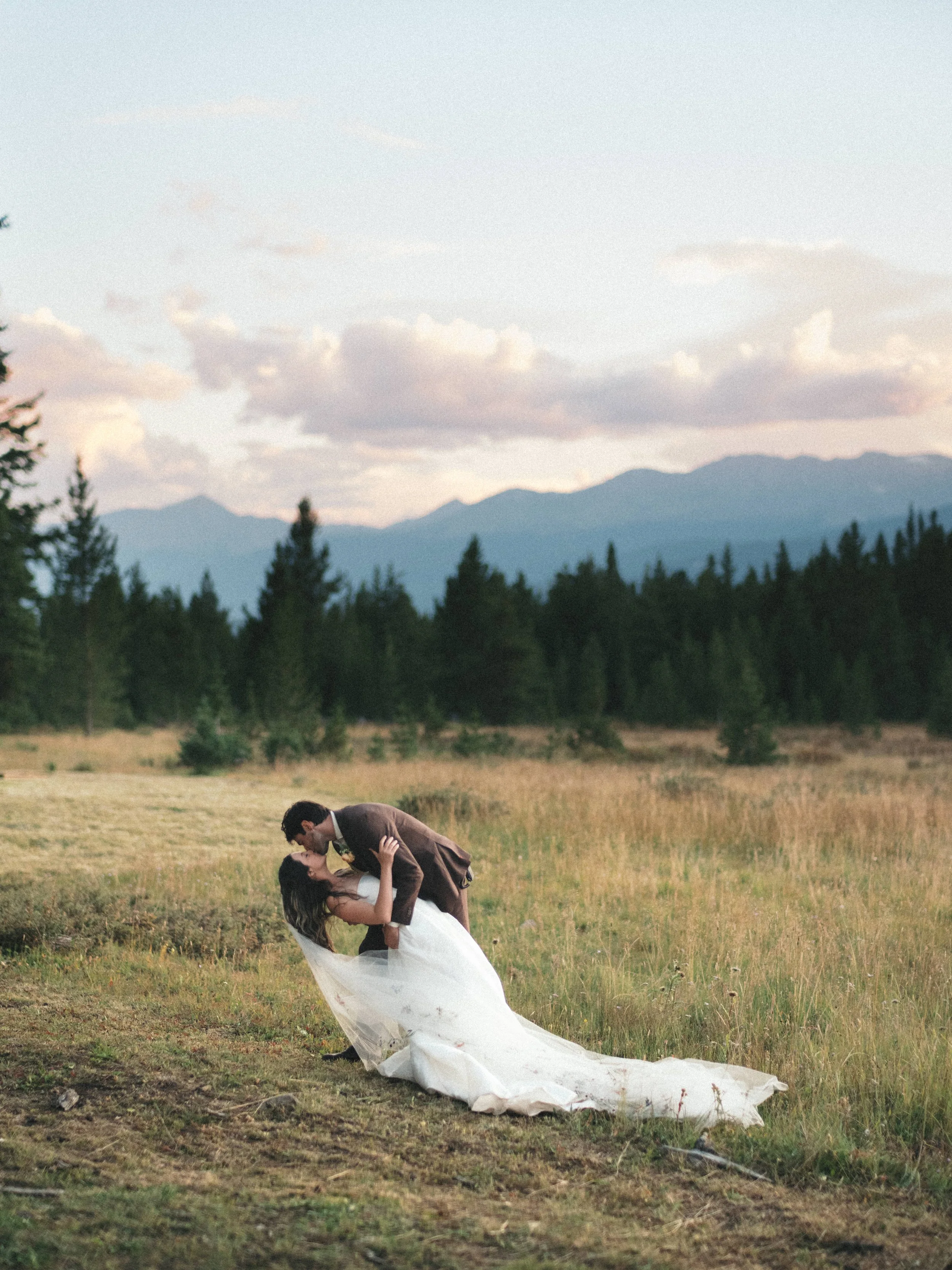 A couple in wedding attire sharing a romantic kiss outdoors in a grassy field with mountains and trees in the background, during sunset or early evening.
