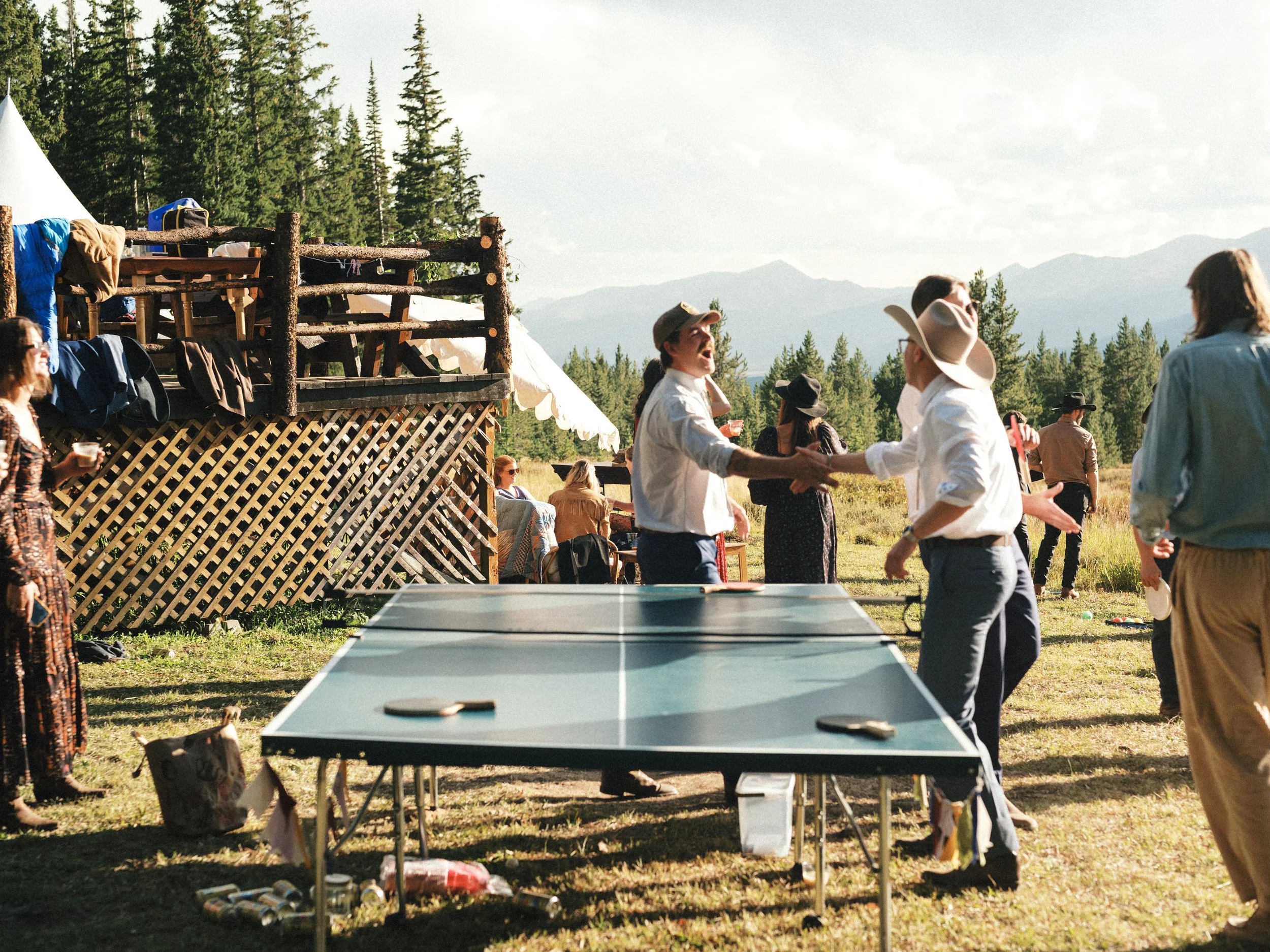 People outdoors at a social gathering near pine trees with mountains in the background, playing ping pong.