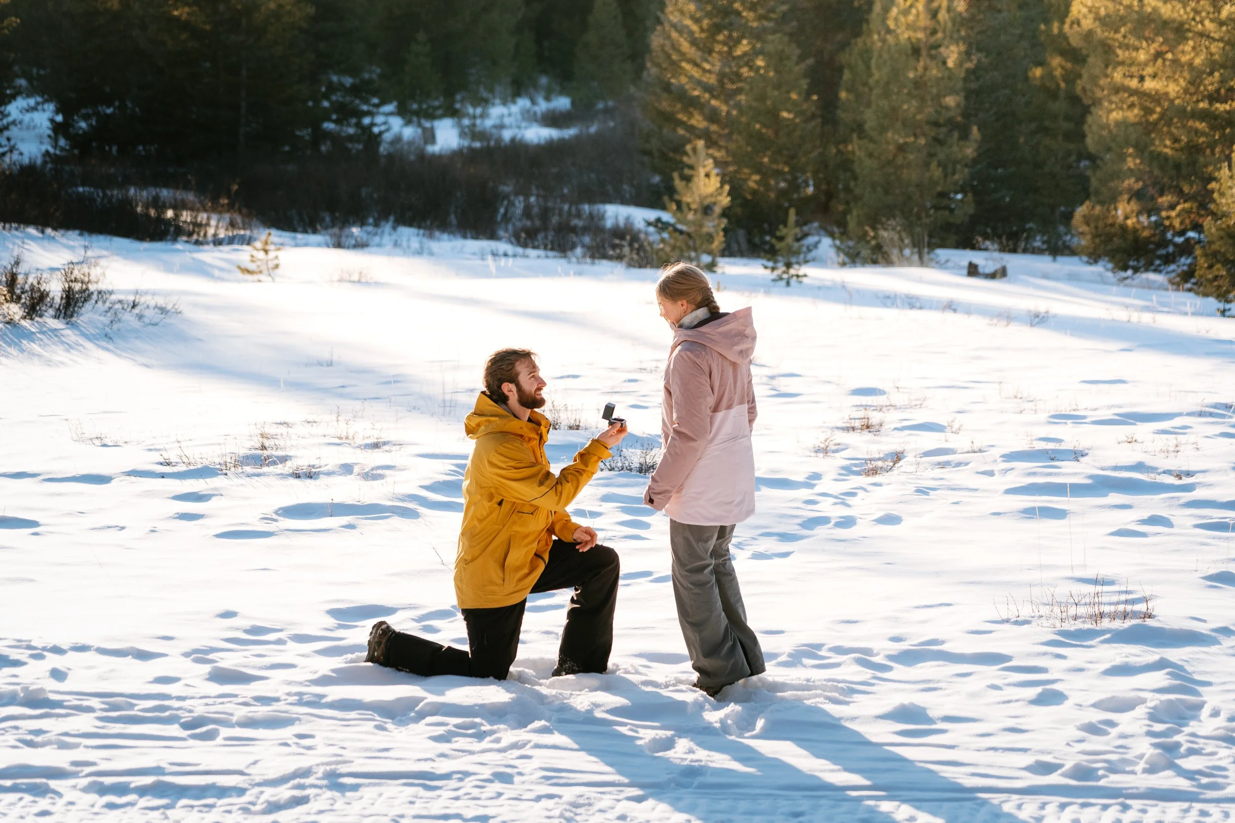 A man proposing marriage to a woman in a snowy outdoor setting with trees in the background.