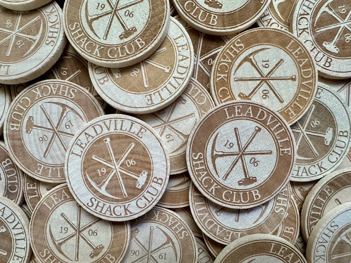 A collection of circular tokens with a wood-like appearance, each featuring the text "Leadville Shack Club" and the year "1906". The tokens have a design with crossed pickaxes and a hammer.