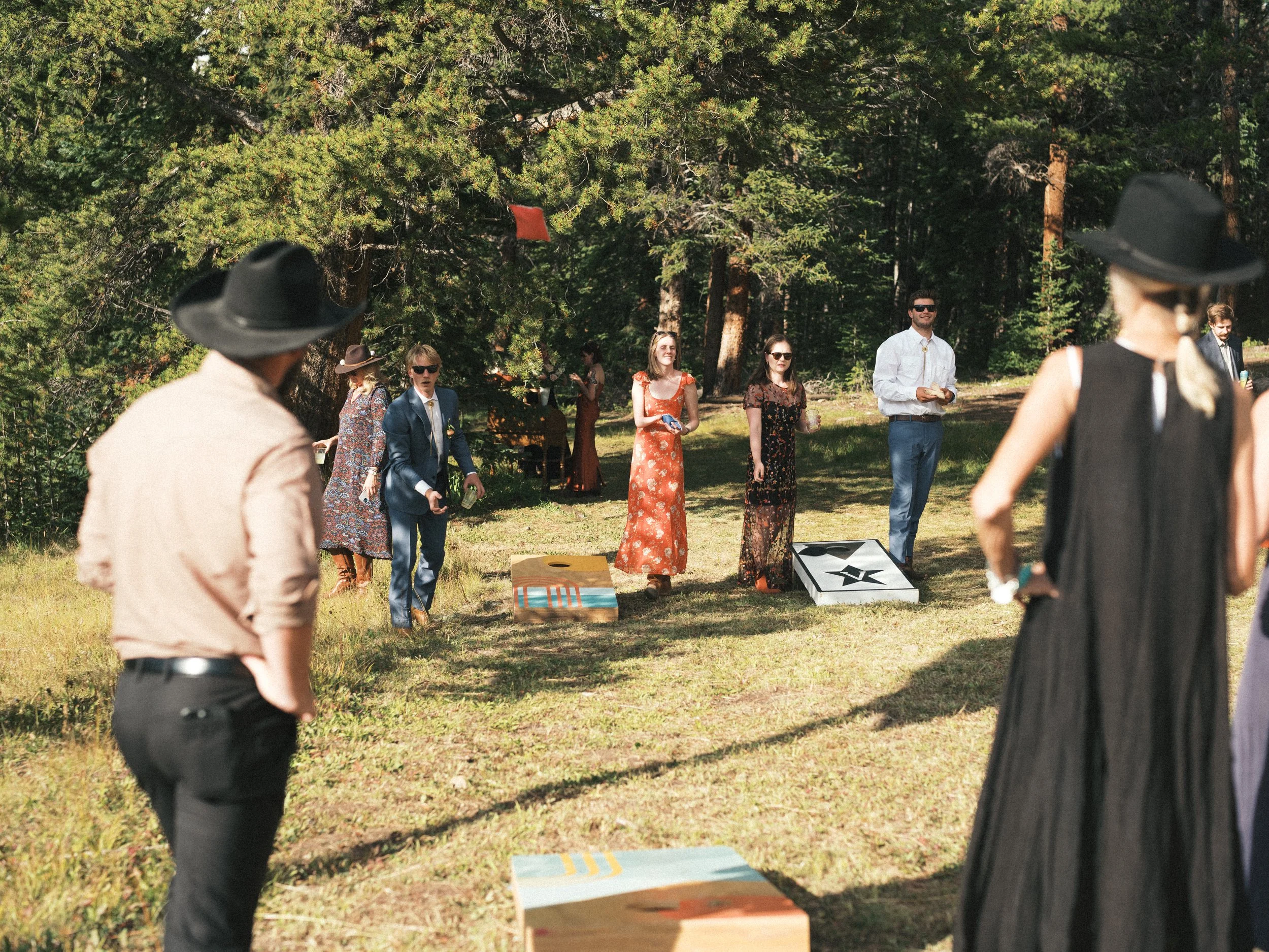 People playing cornhole outdoors in a wooded park, with some wearing cowboy hats and sunglasses, and a red square flag hanging from a tree.