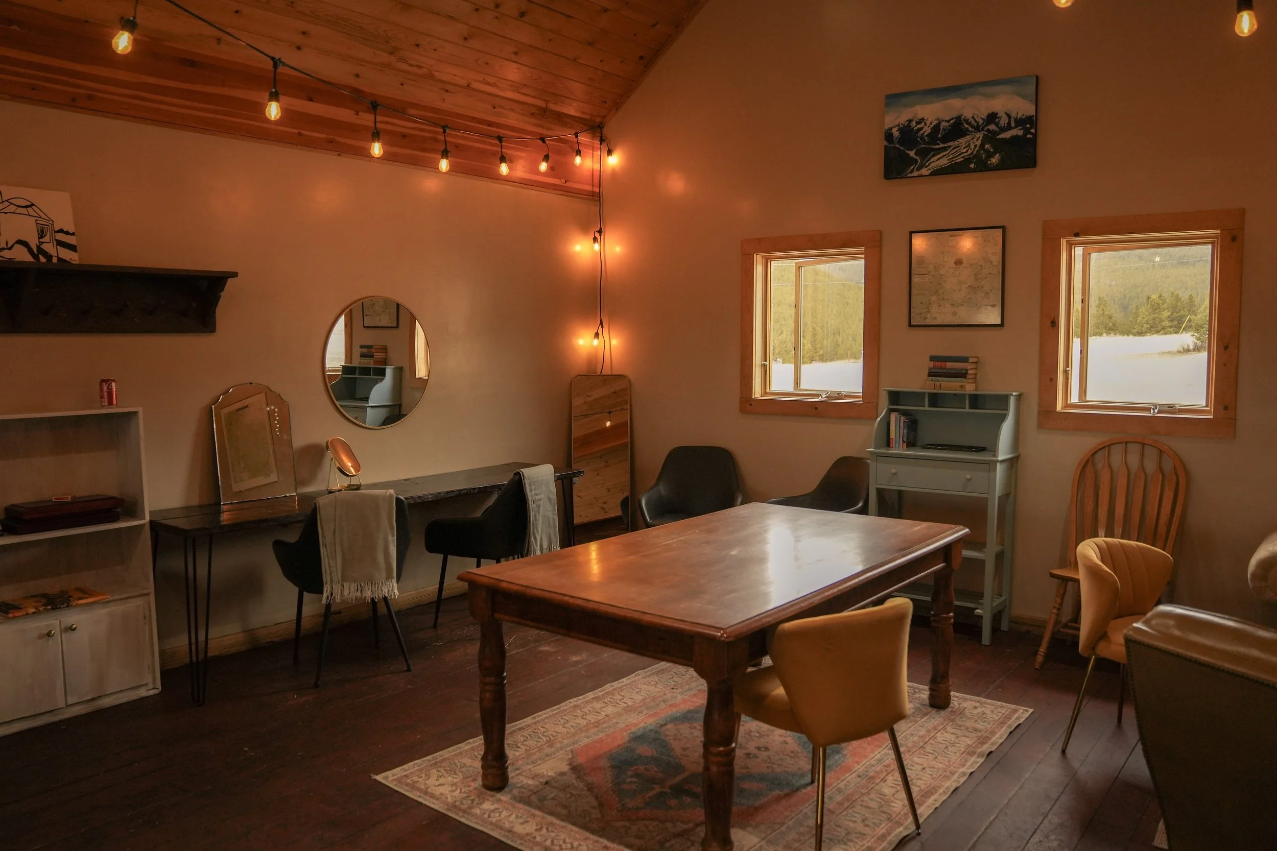 A cozy living room with wooden ceiling and two windows showing an outdoor landscape with trees. The room has string lights hanging from the ceiling, a mirror on the wall, a table surrounded by chairs, and bookshelves with decorative items.