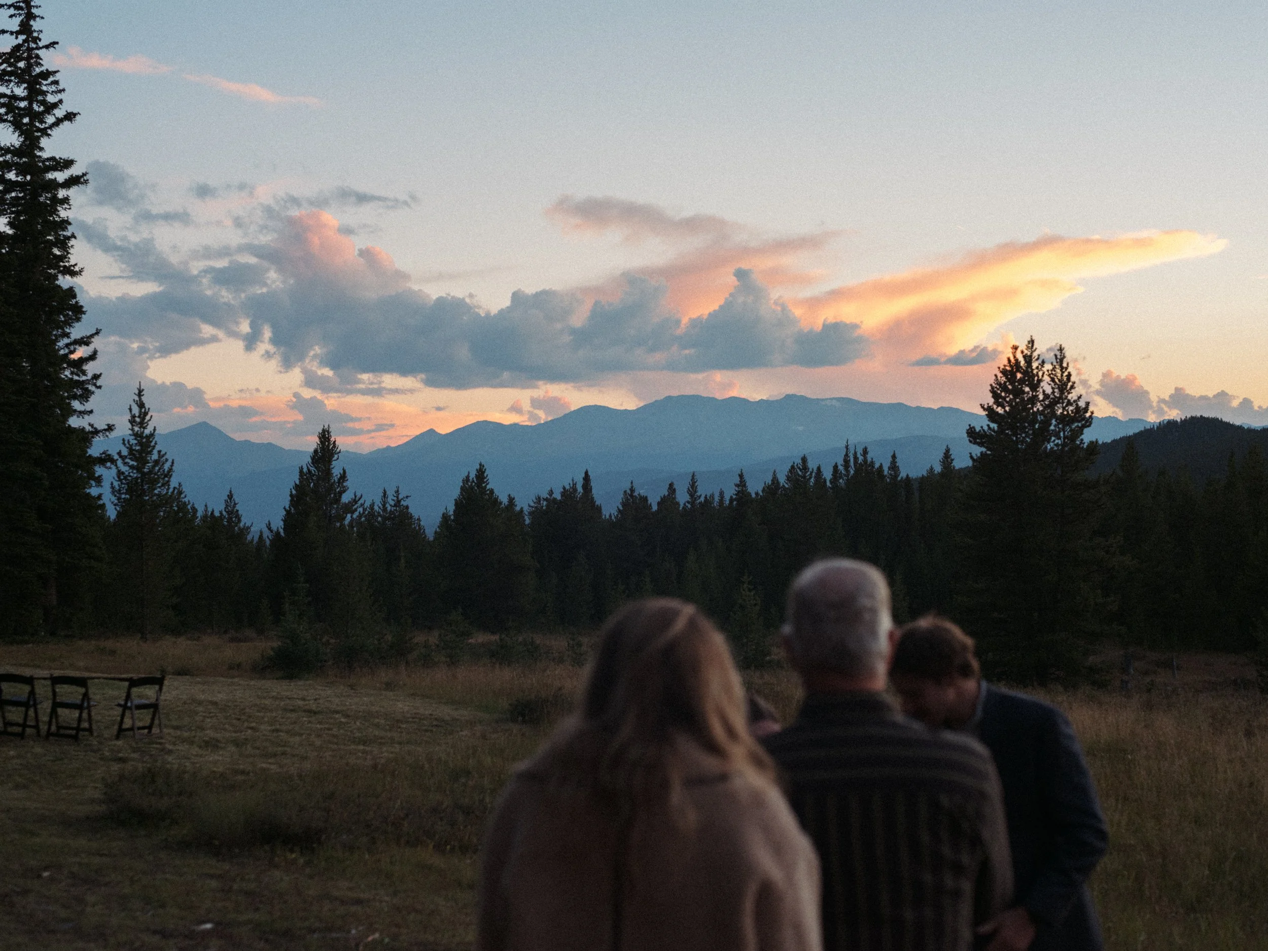 A group of people sitting outdoors on chairs, watching a sunset over mountains and clouds.