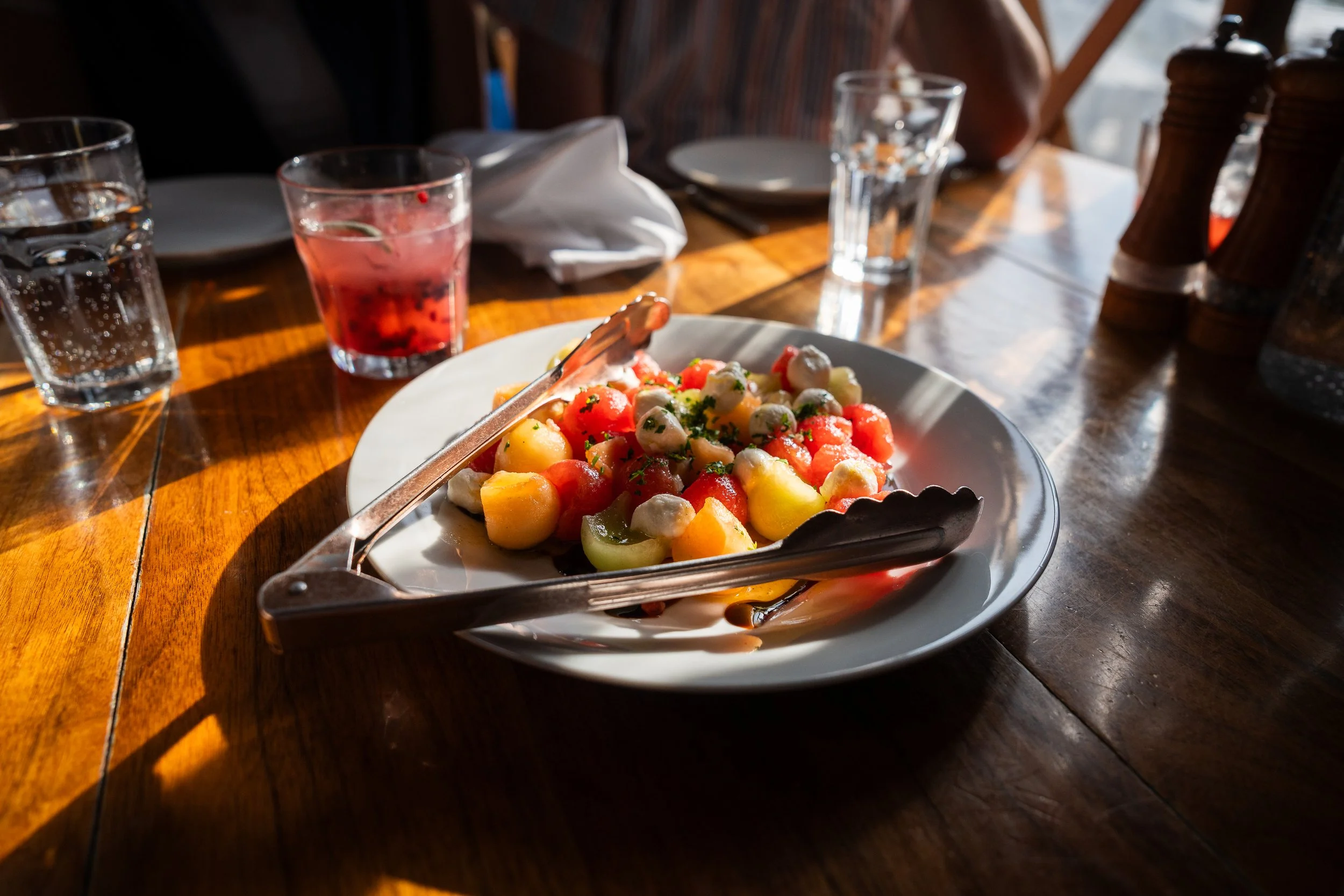 A plate of colorful gnocchi pasta with vegetables, with tongs resting on top. The table has three glasses of water and a glass of pink drink, with salt and pepper shakers nearby.