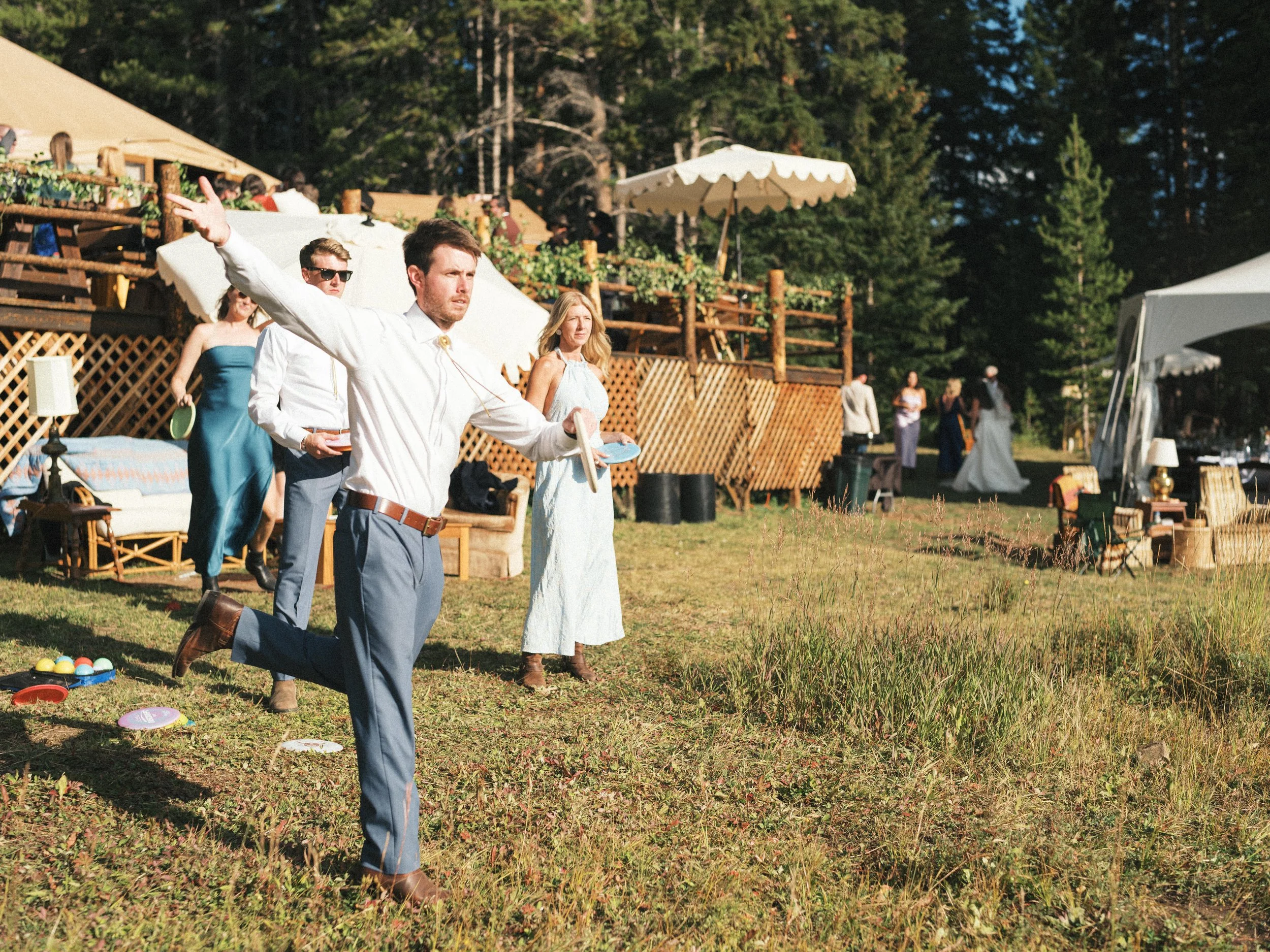 People participating in a game of lawn bowling on a grassy field, with a man in a white shirt holding a blue disc, other players watching, and a backdrop of trees and tents at an outdoor gathering.