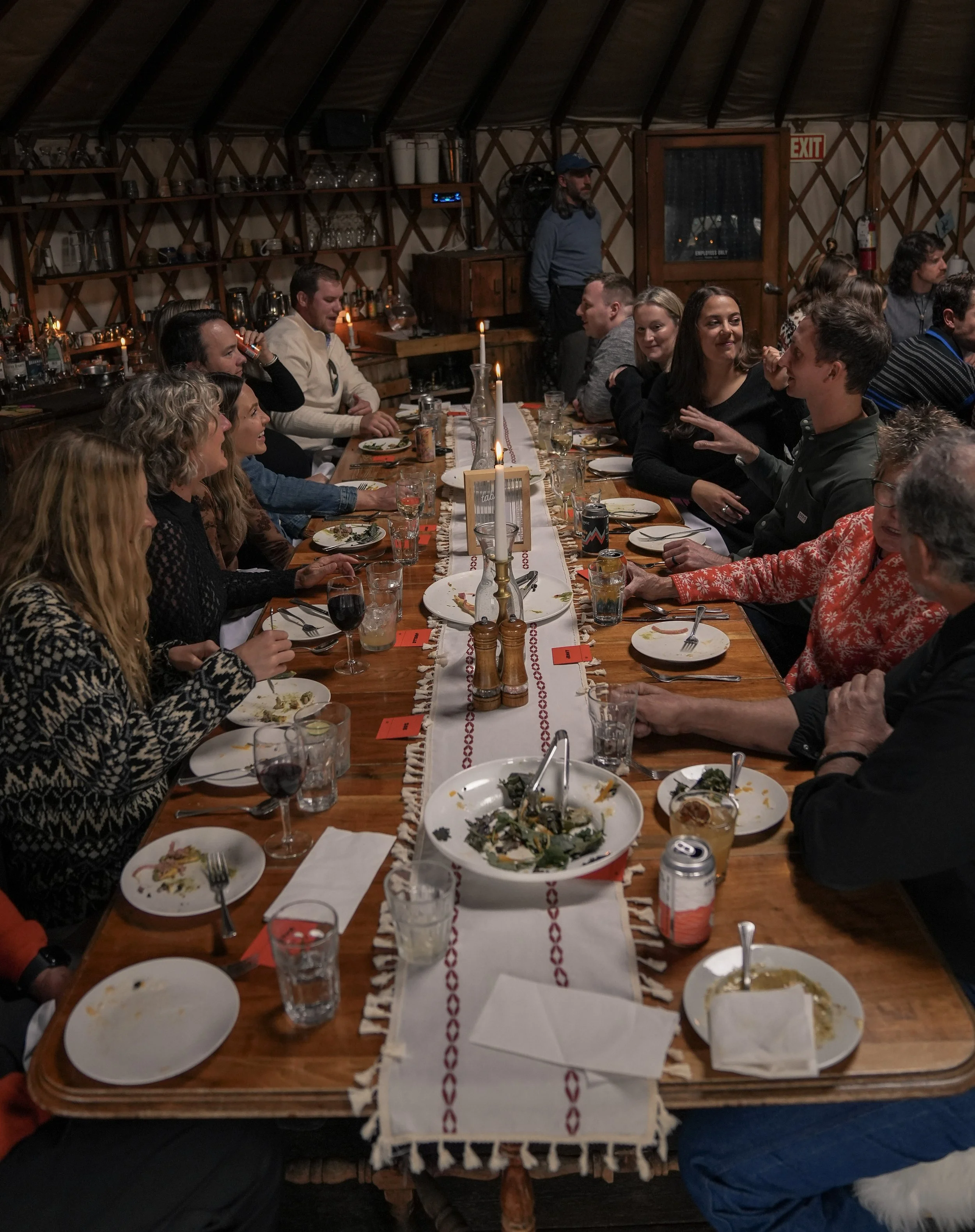 People gathered around a long wooden dining table sharing a meal in a warm, rustic restaurant. The table has a white decorative tablecloth, partially eaten food, wine glasses, and candles. The background shows shelves with glasses and bottles, and a 