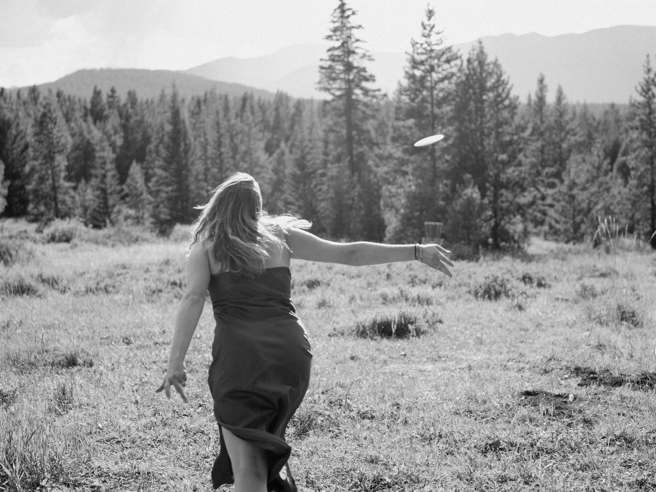 A woman playing frisbee outdoors in a grassy field with trees and mountains in the background.