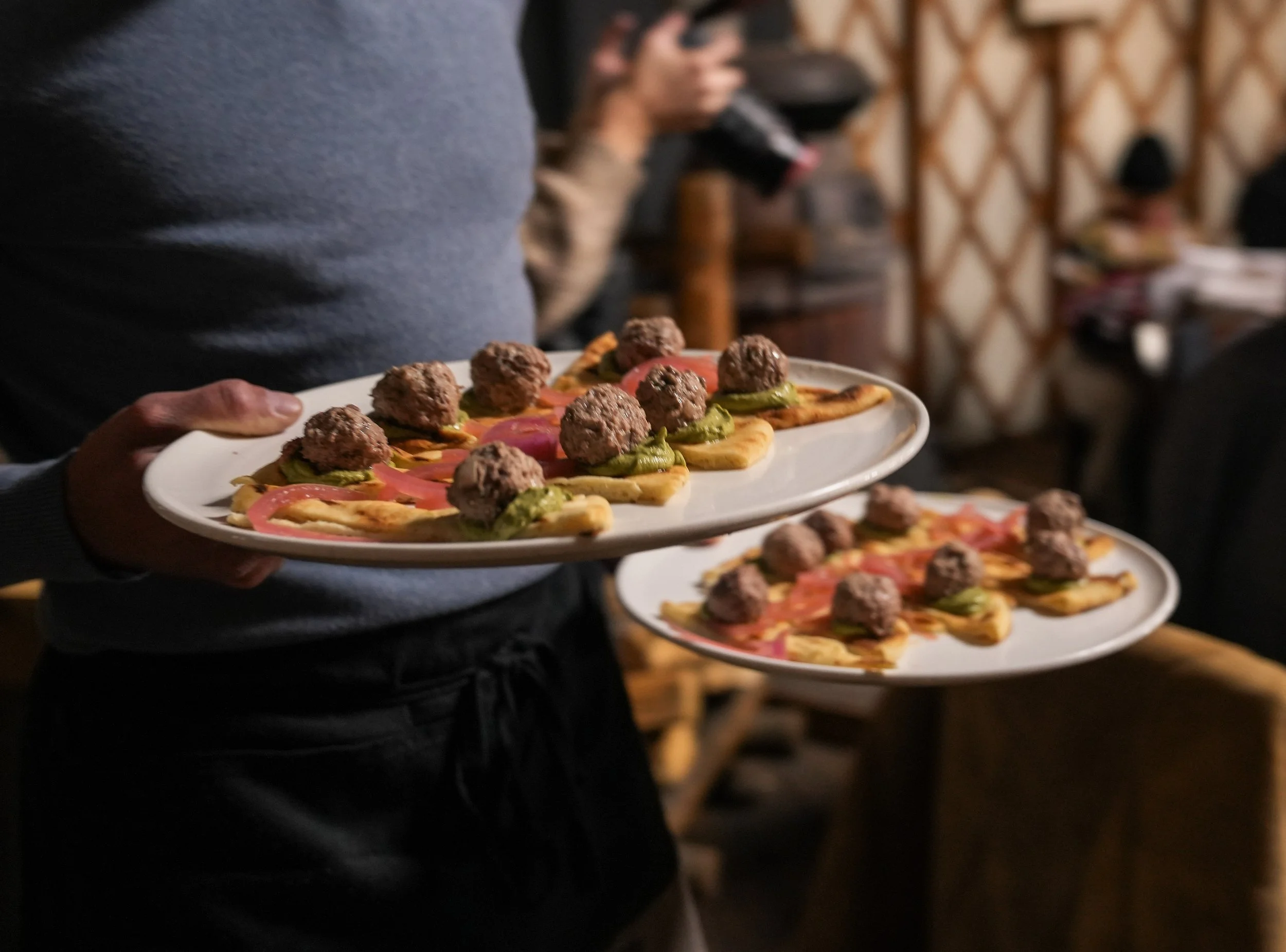 A person is holding two plates of food, each topped with meatballs, slices of tomato, and green peppers, with a blurred background of a restaurant setting.