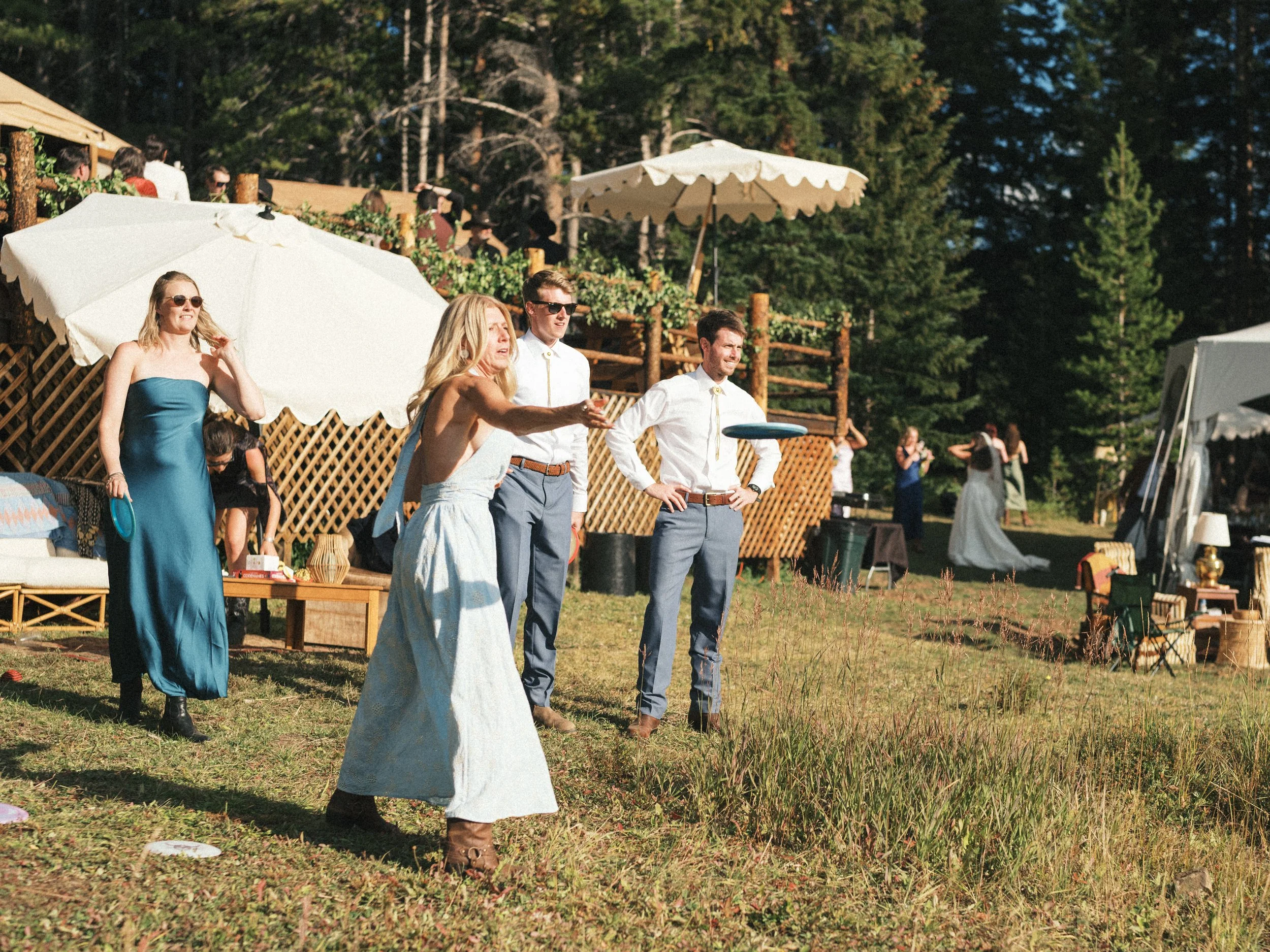 People standing and socializing outdoors at a daytime event with tents and umbrellas, surrounded by trees.