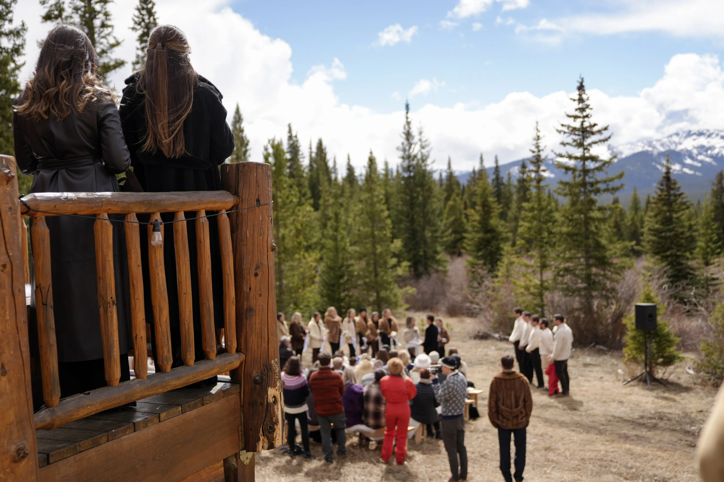 Two women stand on a wooden platform overlooking an outdoor ceremony with people dressed in formal attire, with a background of pine trees, mountains, and a cloudy sky.