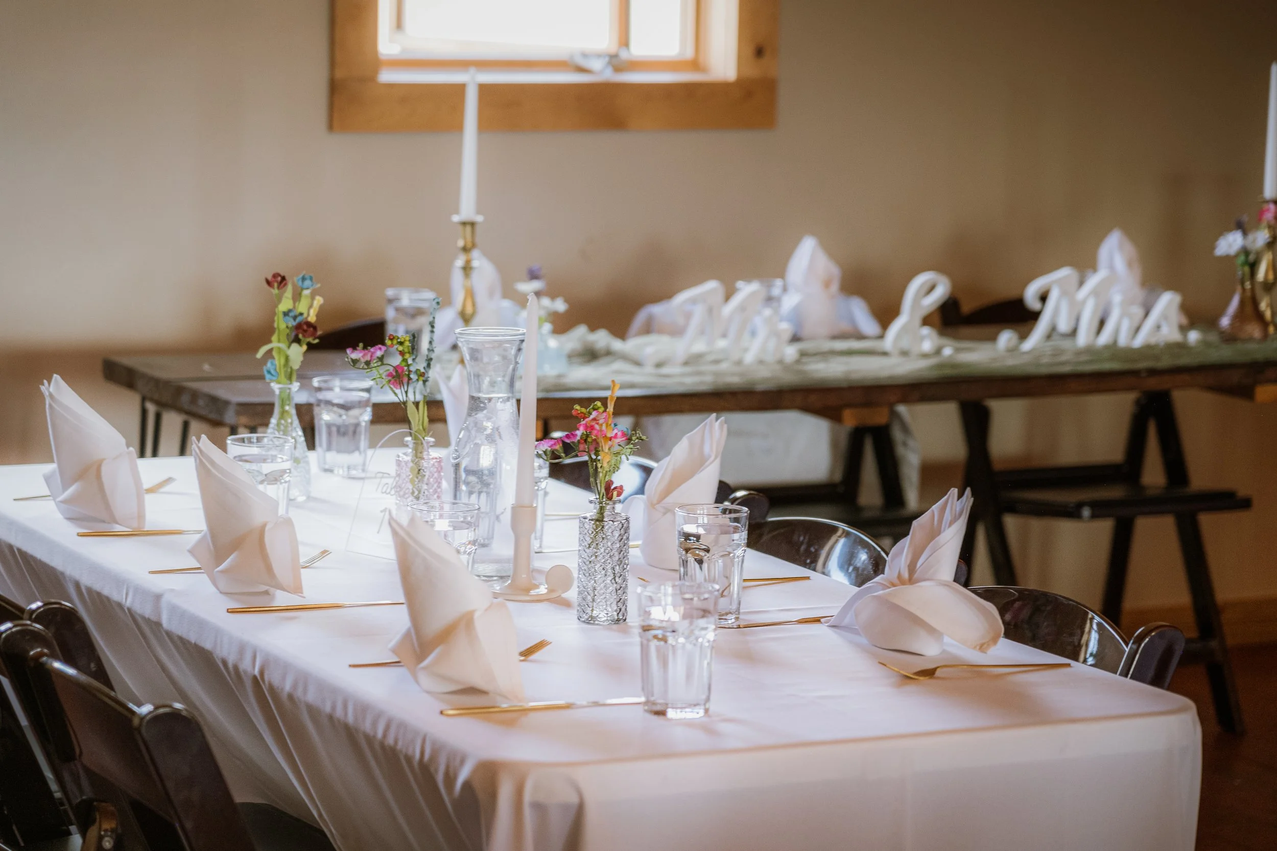 Decorated banquet table set for a formal event with white napkins, glassware, and small colorful flower arrangements, with a decorative wooden sign in the background.
