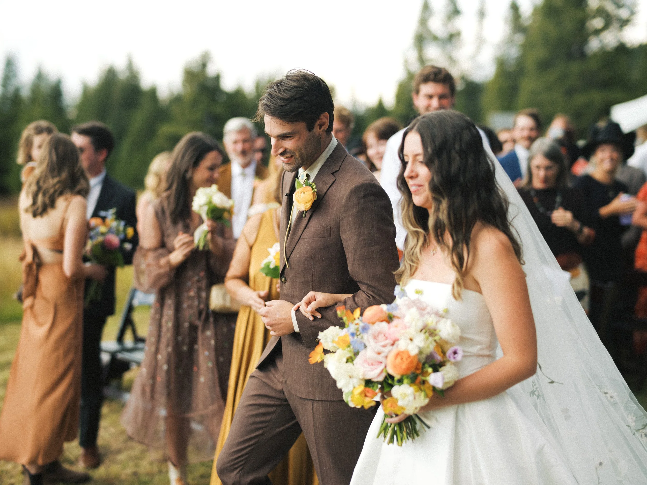 A bride and groom in wedding attire surrounded by smiling guests outdoors, with trees in the background.
