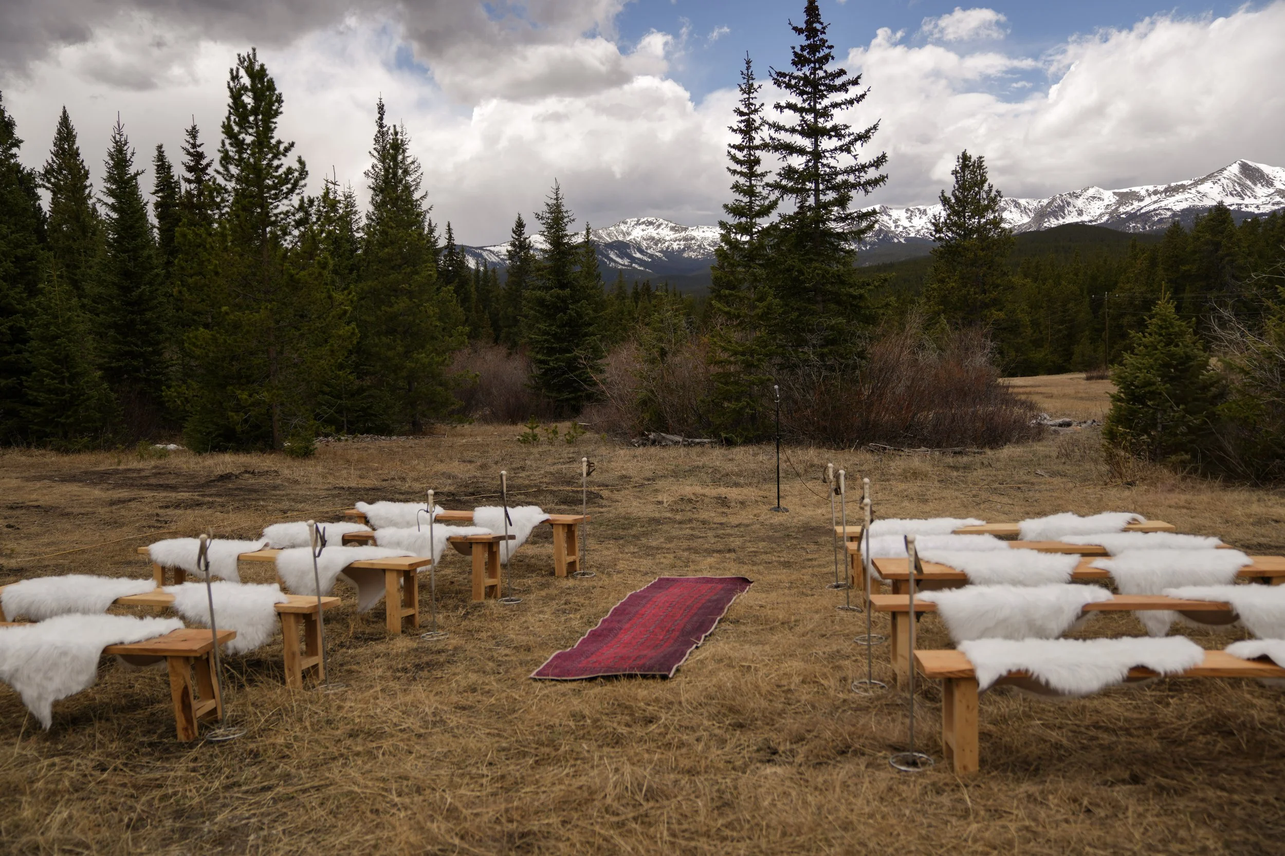 Outdoor setting with wooden benches covered in white sheepskin, arranged in rows on a grassy field, with a red carpet walkway in the center. In the background, there are tall evergreen trees and snow-capped mountains beneath a partly cloudy sky.
