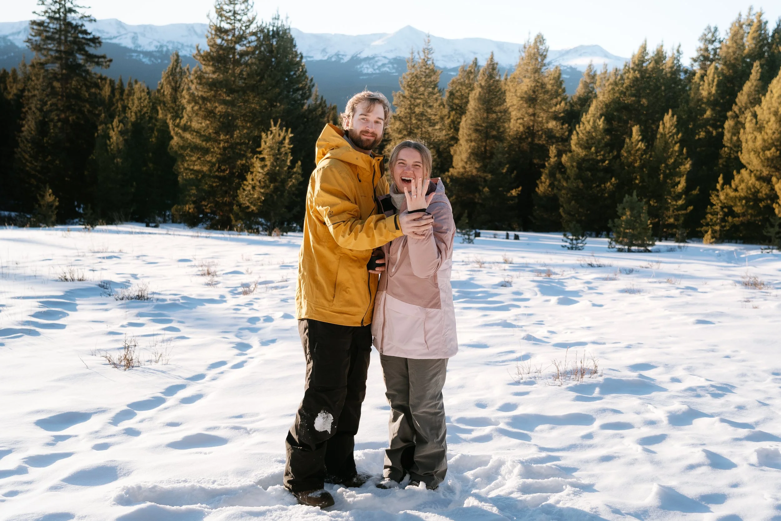 A man and a woman standing together in a snowy field of a forest with mountains in the background. Both are smiling and wearing winter jackets and snow boots. The woman is showing her hand with a ring, indicating they are engaged or married.