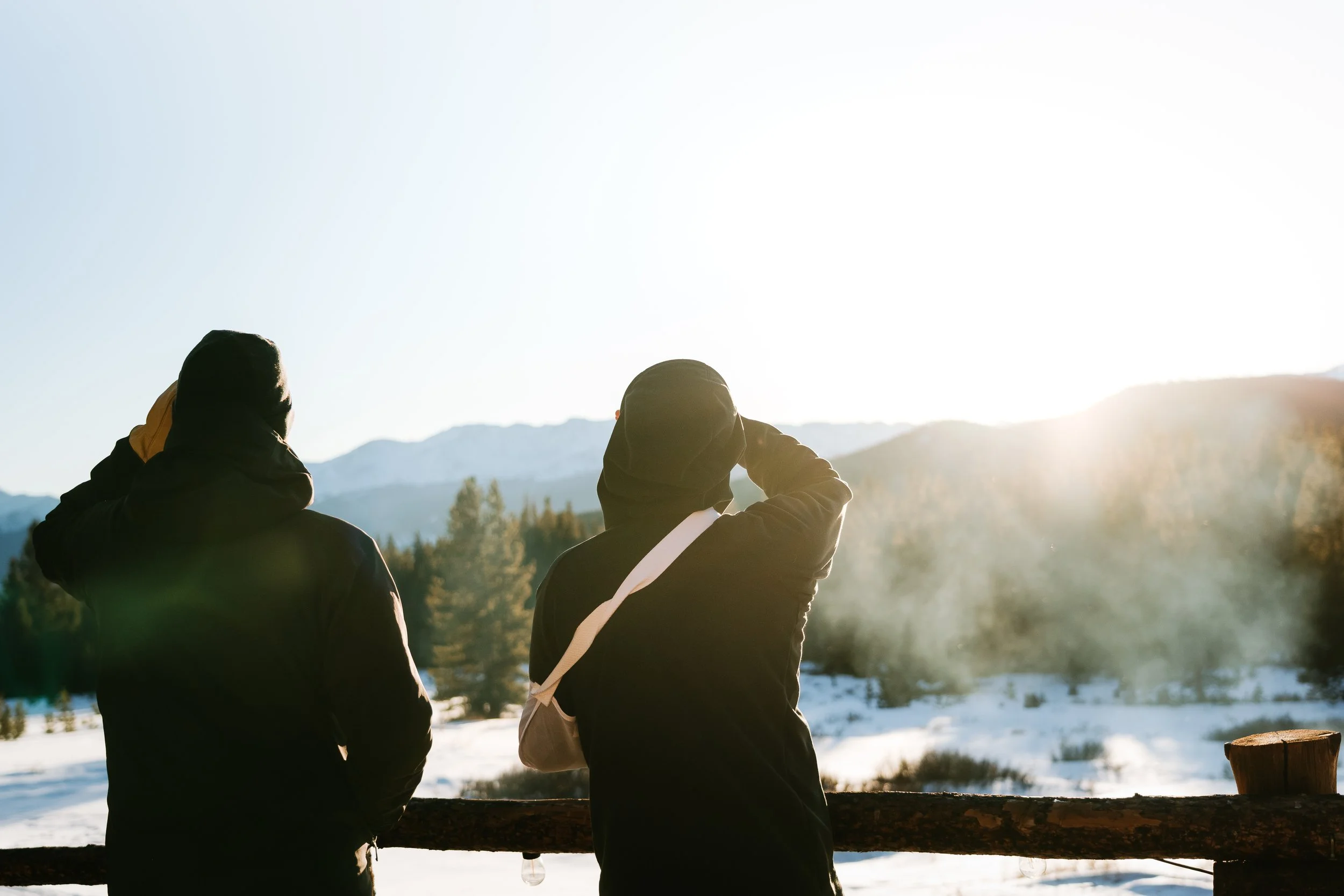 Three people wearing hooded jackets standing outdoors in a snowy landscape with mountains in the background, backlit by the sun.