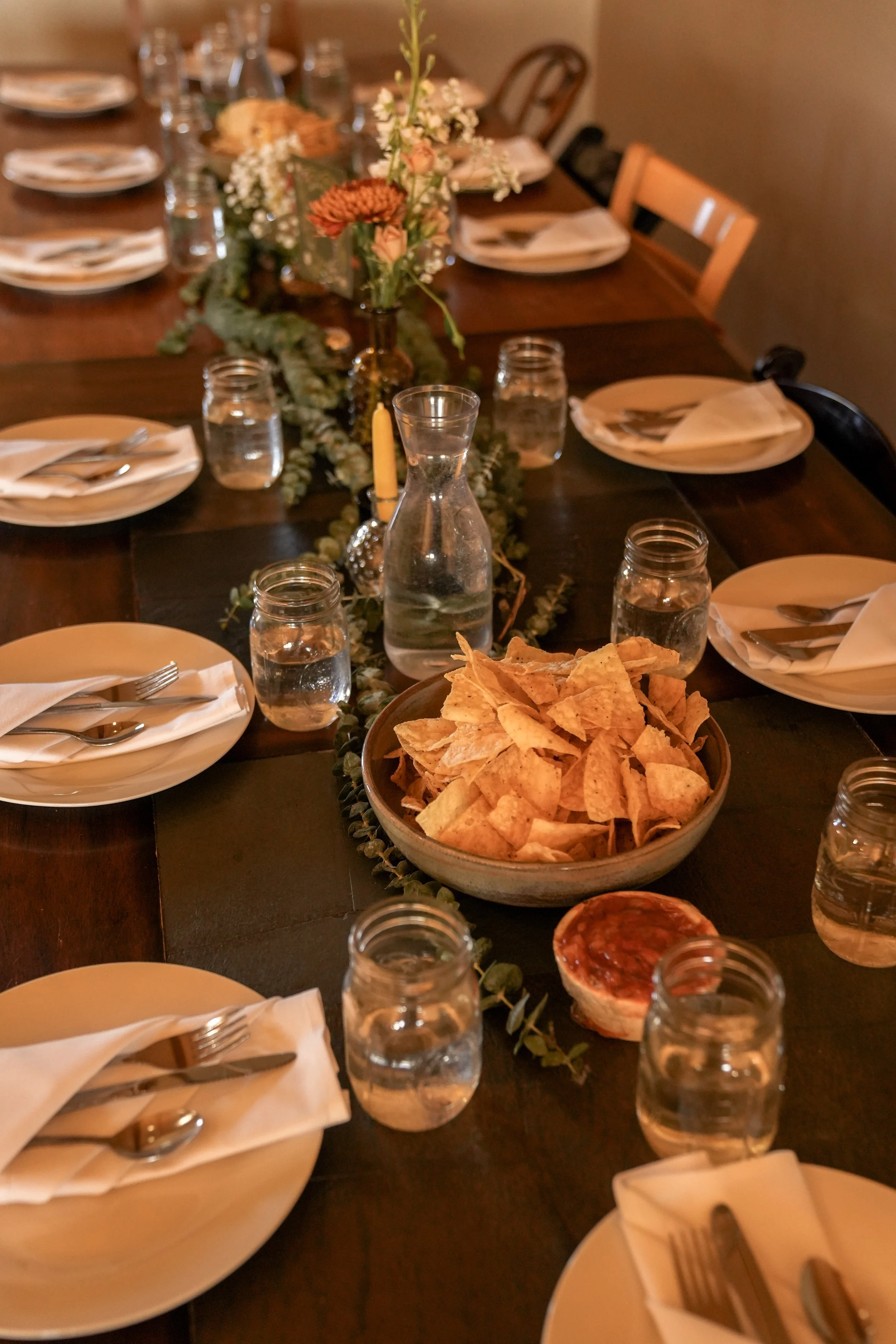 A dinner table set with plates, napkins, silverware, glasses of water, a centerpiece with flowers, a jar candle, a pitcher of water, and a bowl of tortilla chips.
