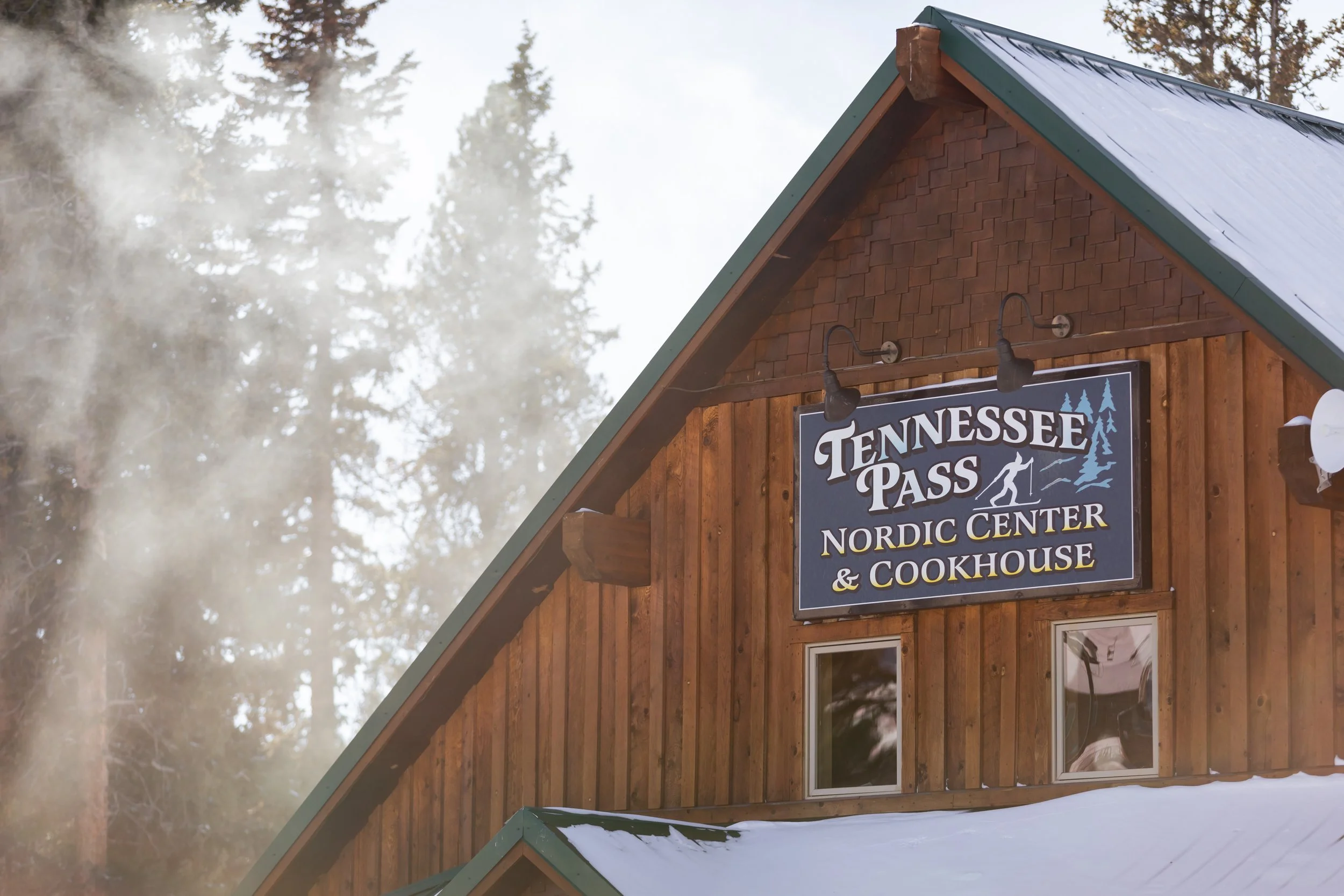 A wooden building with a sign reading "Tennessee Pass Nordic Center & Cookhouse" and a snow-covered roof, surrounded by trees and misty weather.