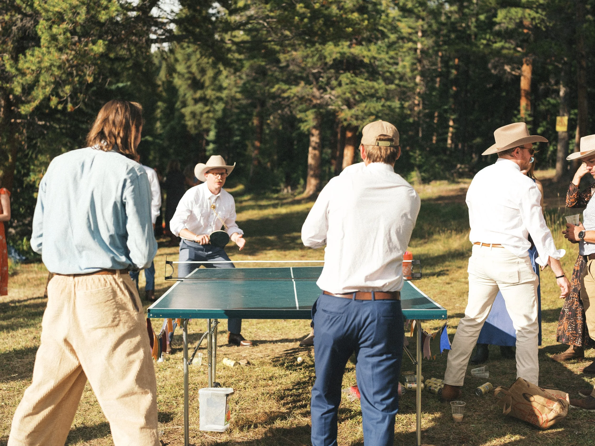 People gathered outdoors playing ping pong in a park with trees in the background, sunlight, and casual summer clothing.