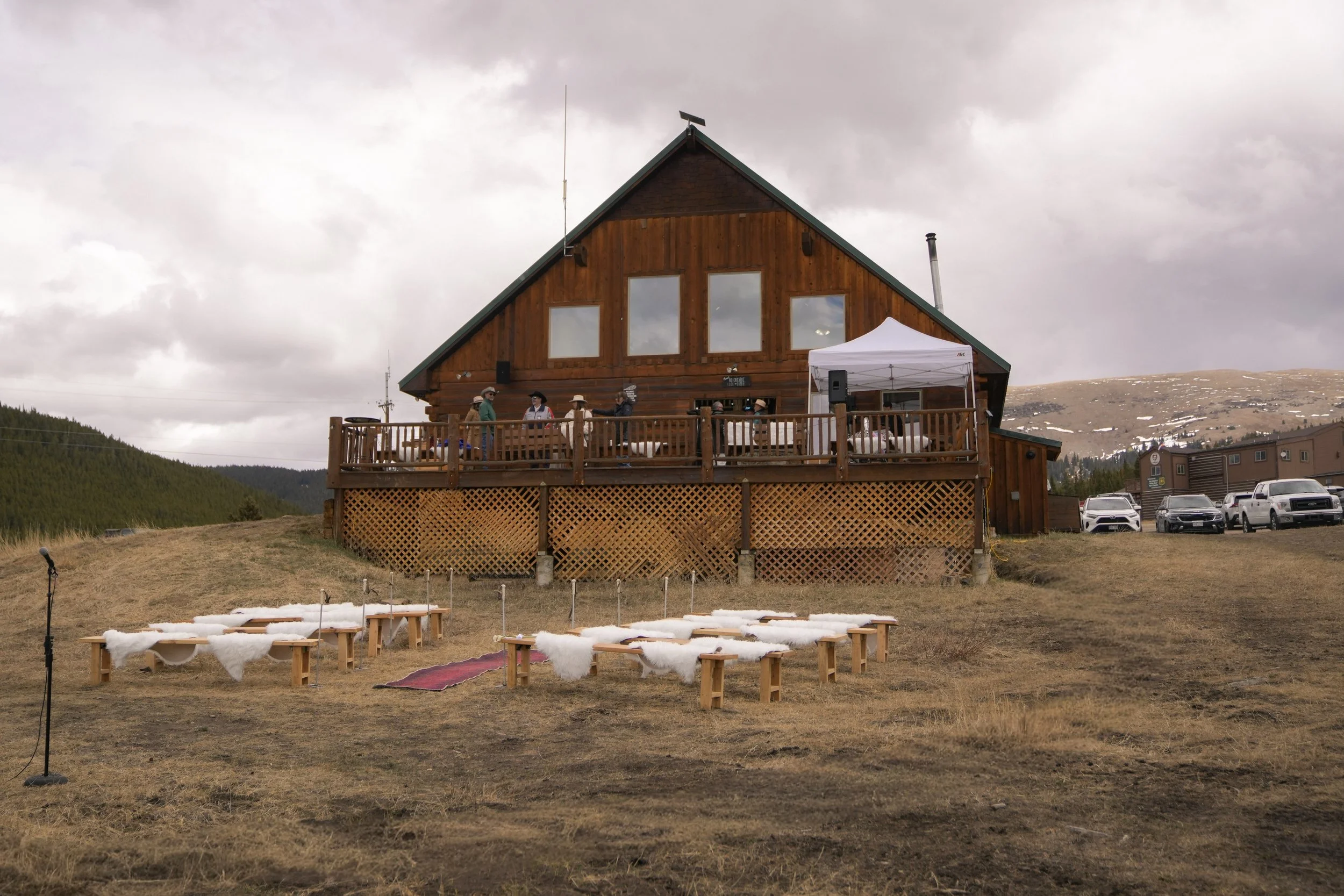 Outdoor wooden cabin on a grassy hill with a deck and a white canopy. Several people are standing and sitting on the deck, and there are chairs arranged on the ground facing the cabin. Cars are parked nearby, and mountains are visible in the backgrou
