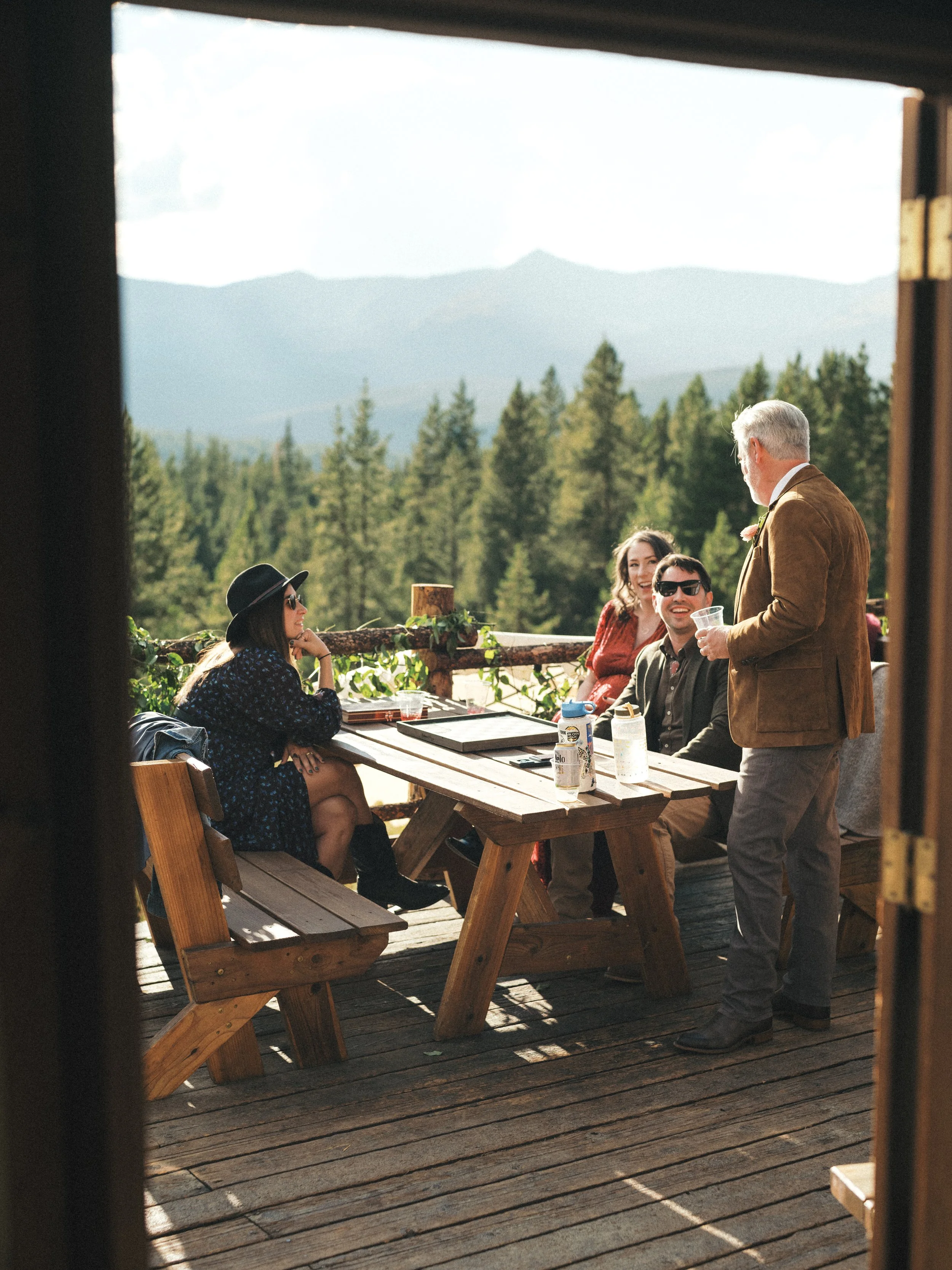 People gathered outdoors at a wooden deck with a mountain view, engaged in conversation, with trees and mountains in the background.