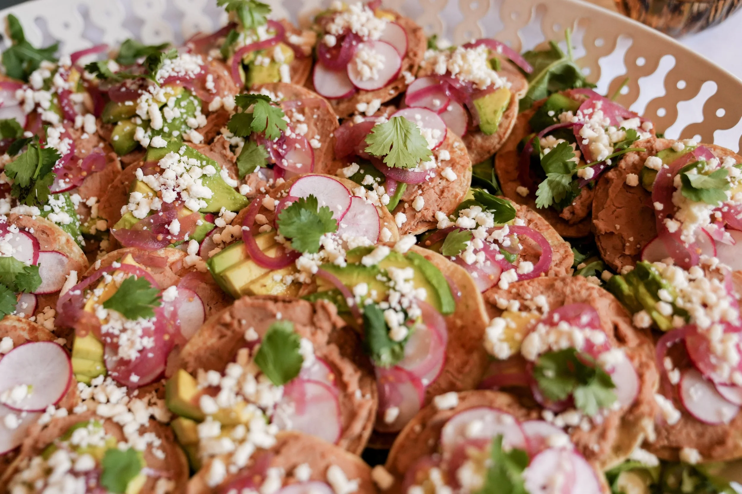 Close-up of multiple small tacos topped with radish slices, avocado, crumbled cheese, cilantro, and pink pickled onions on a tray.