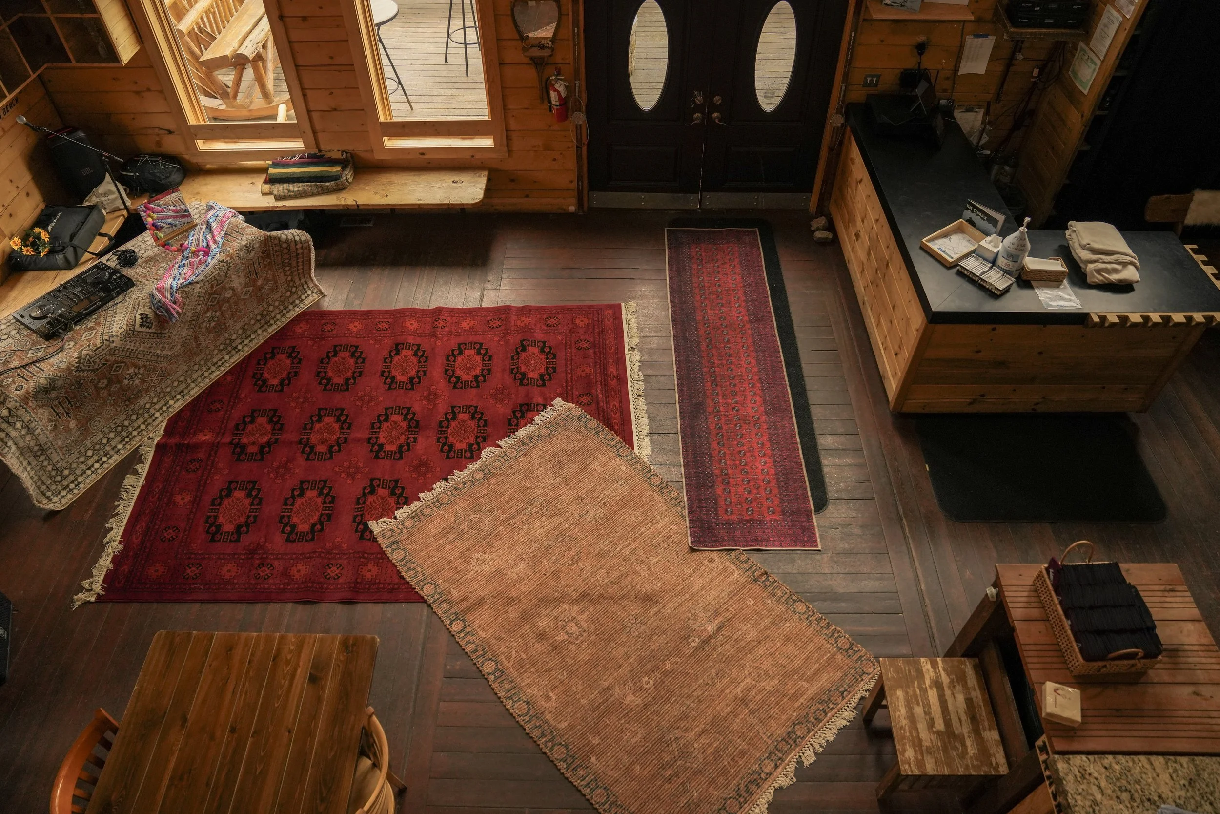 Interior view of a wooden cabin with a black door, multiple red and beige rugs on a wooden floor, a counter with various items, and a window showing an outdoor deck.