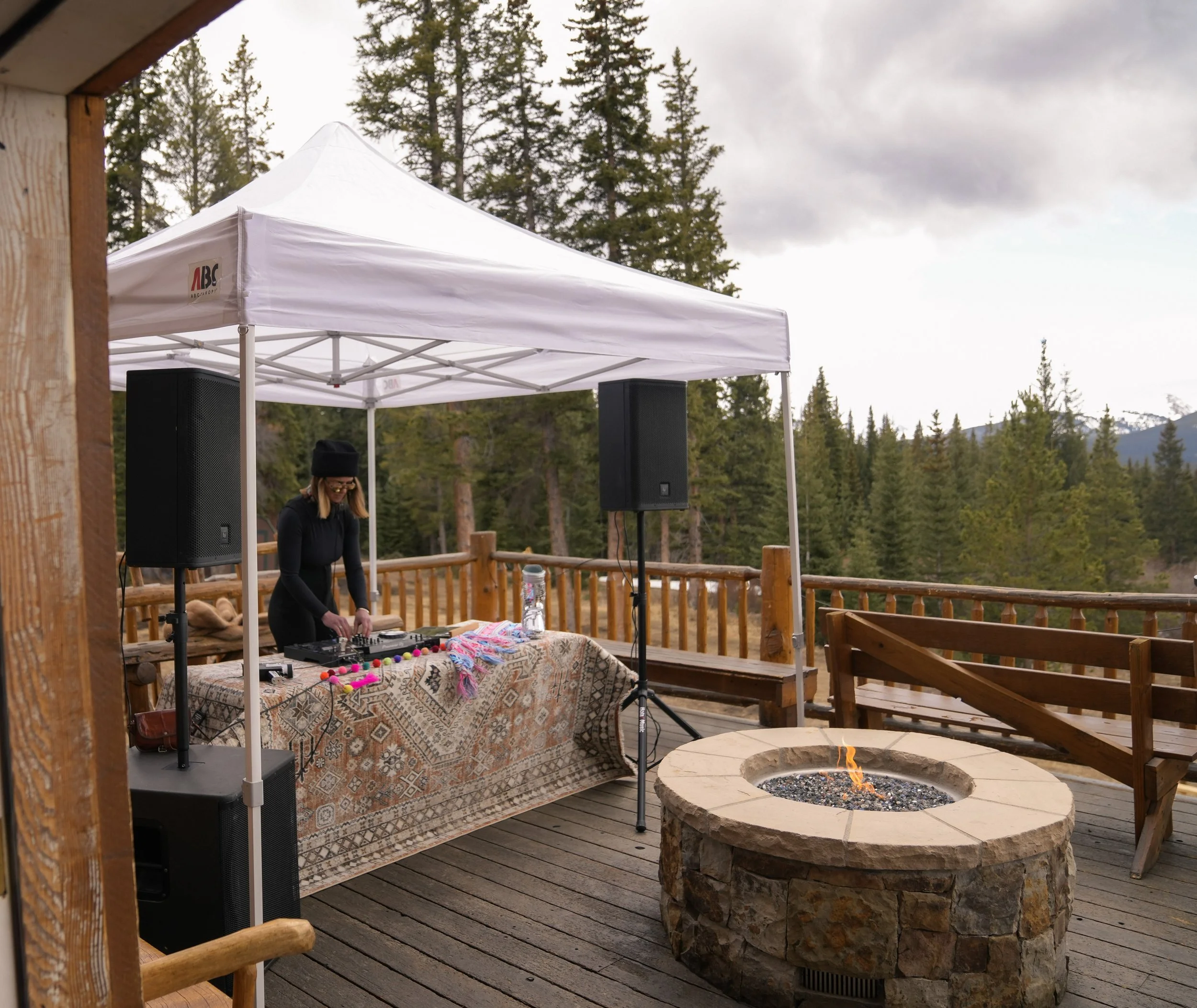 Outdoor event space with a white canopy tent, black speakers, a woman with headphones, decorated table, a fire pit, and trees in the background.