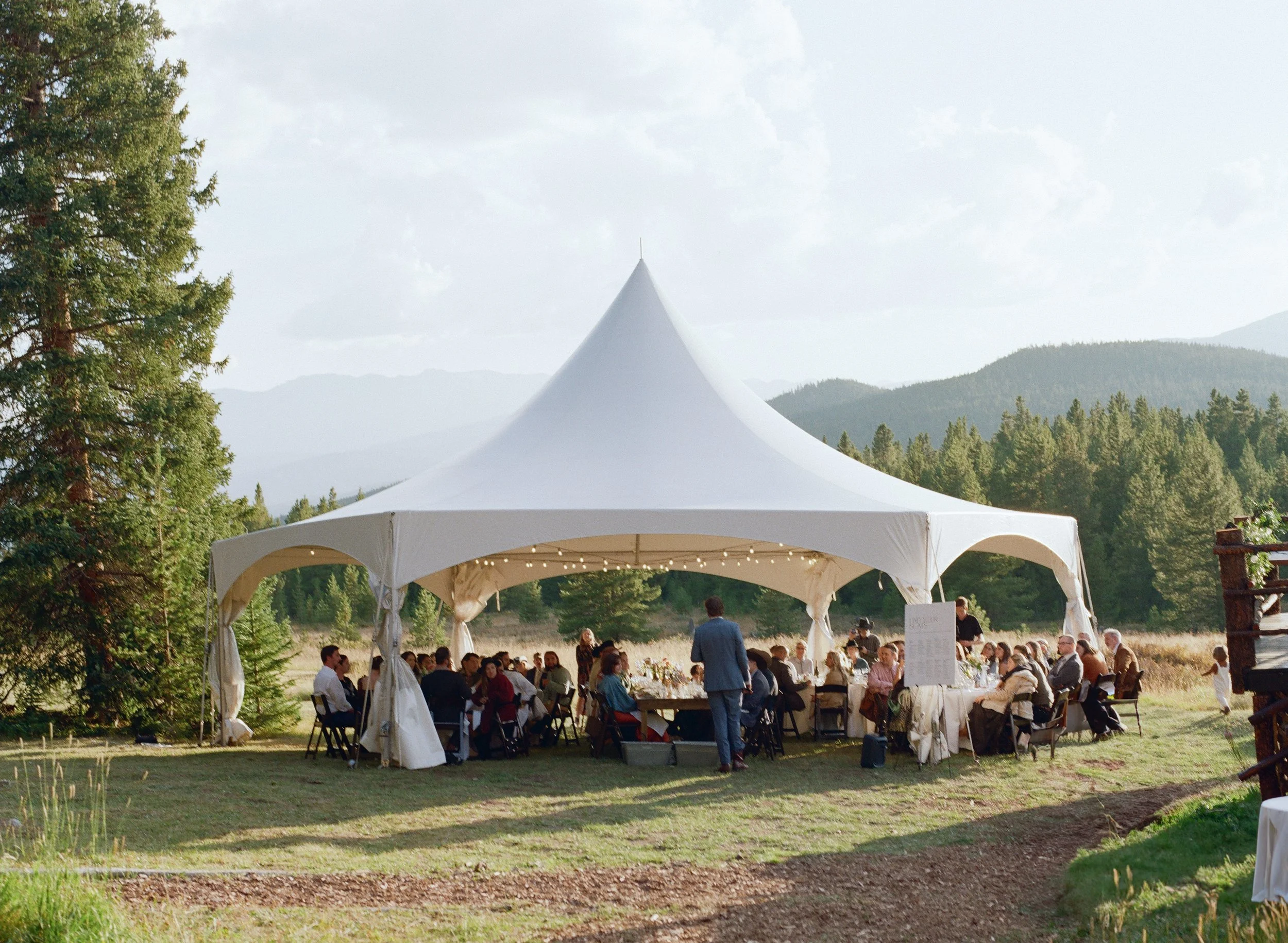 A white event tent set up in an outdoor area with people seated inside, surrounded by trees and mountains in the background, during daytime.