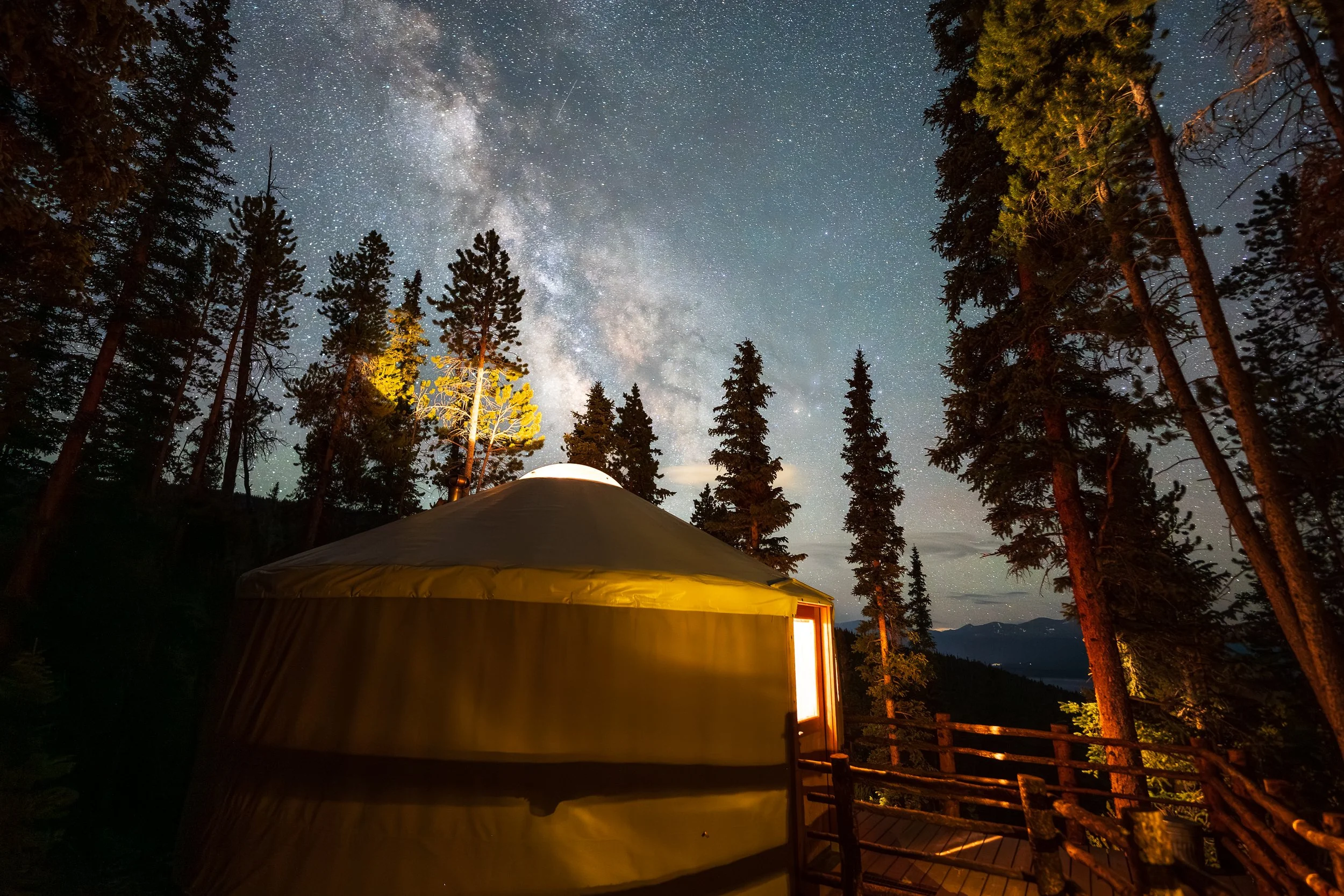 A night sky filled with stars and the Milky Way galaxy seen through a forest of tall pin trees. In the foreground, there is a rounded structure with a window emitting warm light, surrounded by a wooden railing.