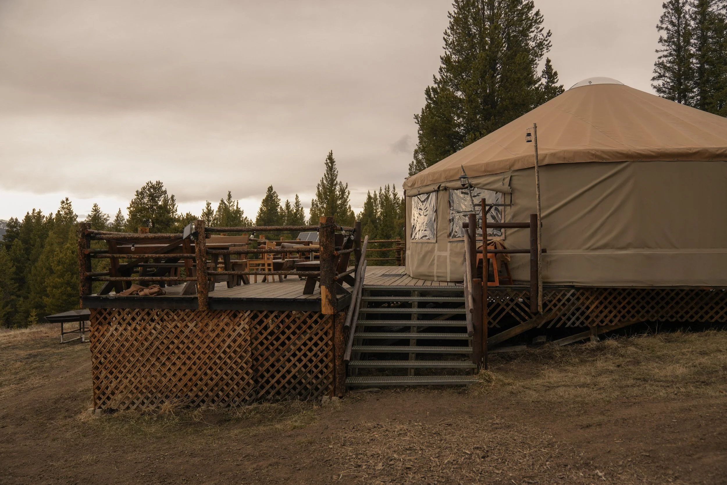 A large beige teepee tent set up on a wooden deck in a forested area with tall pine trees, cloudy sky, and outdoor furniture surrounding it.
