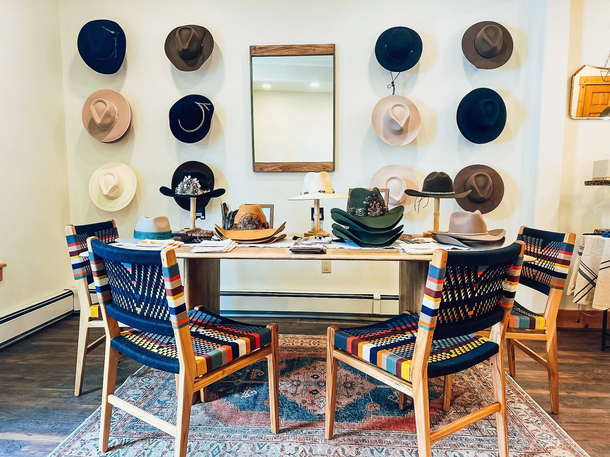 A display of various colored hats hanging on a white wall and seated colorful chairs around a wooden table with hats and catalogs inside, situated in a room with hardwood floors and a mirror.
