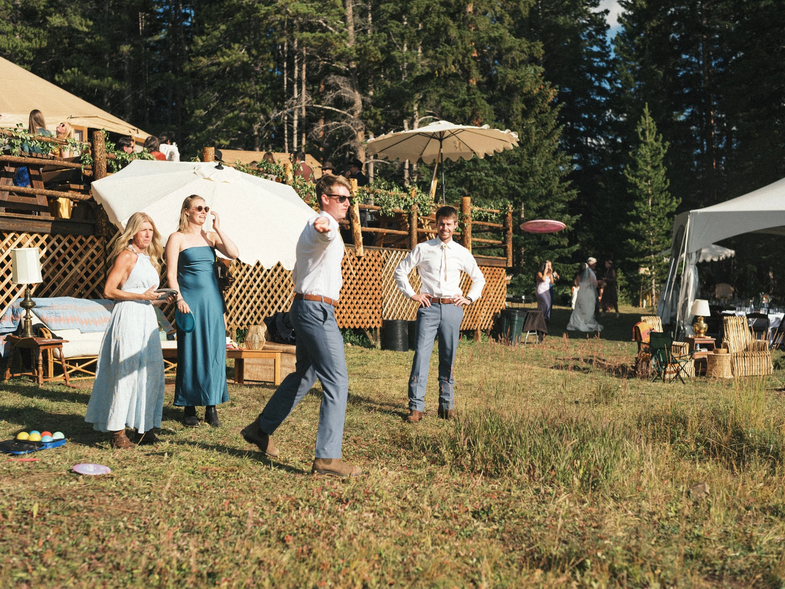 People playing cornhole outside at a yard party, with a wooded area in the background, using umbrellas for shade.