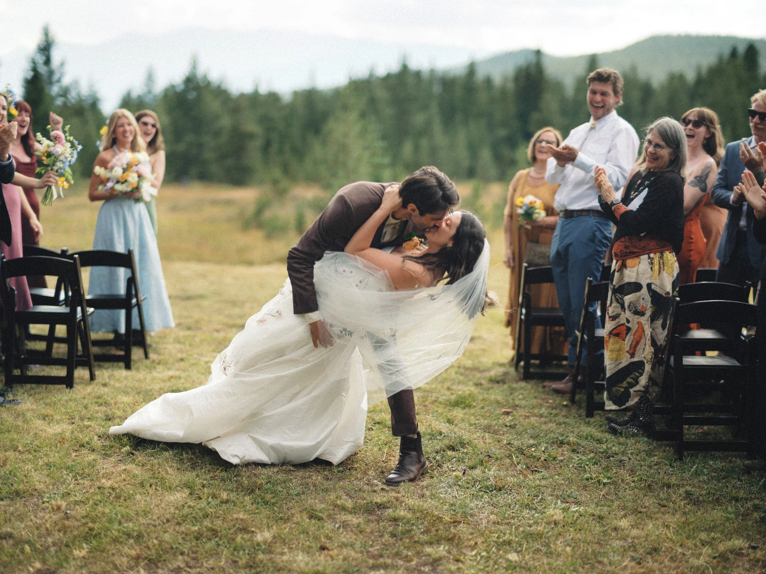 A newlywed couple sharing a kiss during an outdoor wedding ceremony, with guests standing and cheering around them on a grassy field with trees and mountains in the background.